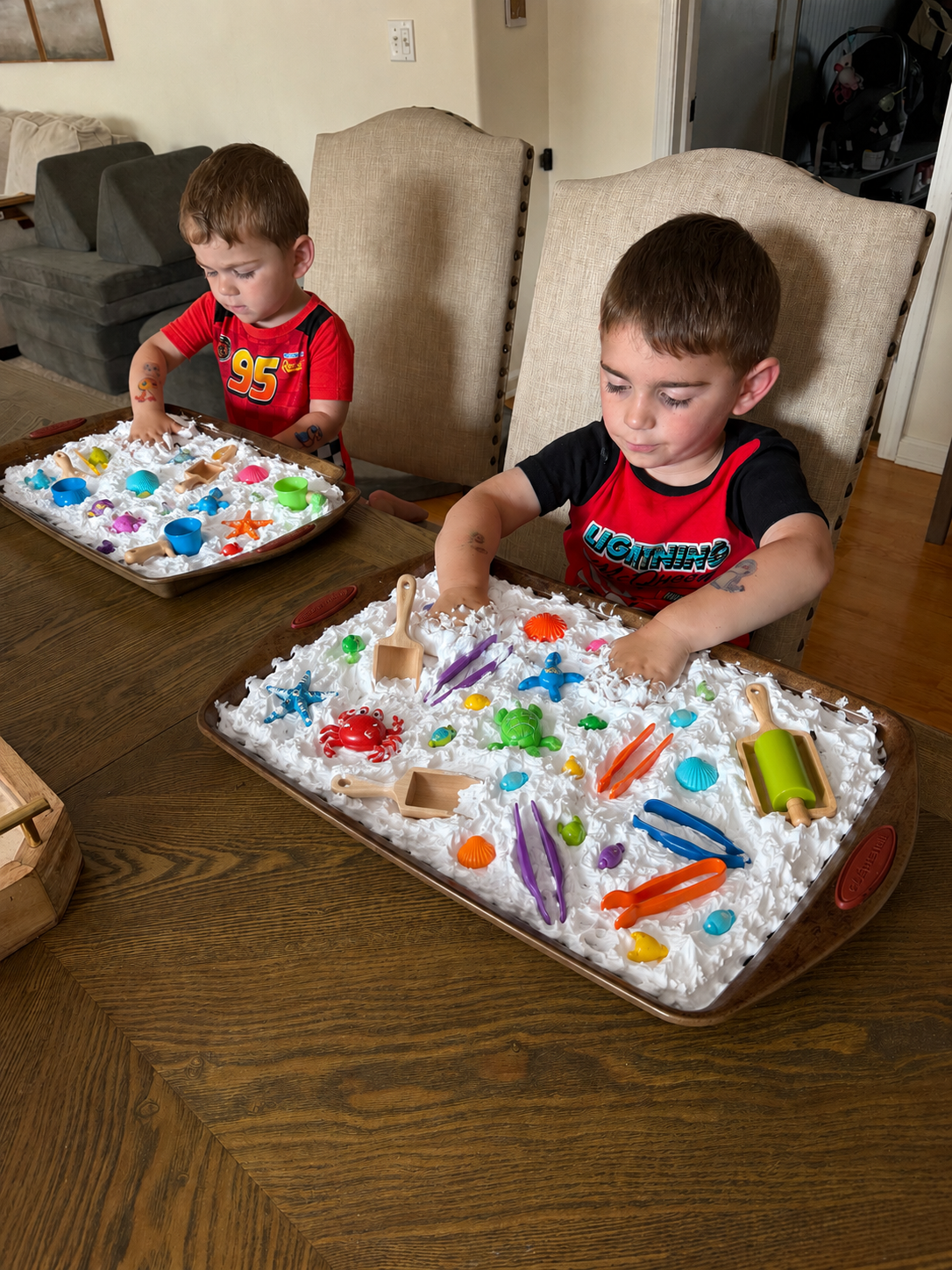 Two young boys playing with sensory bin filled with white foam and miniature beach toys on a wooden table in a dining room.