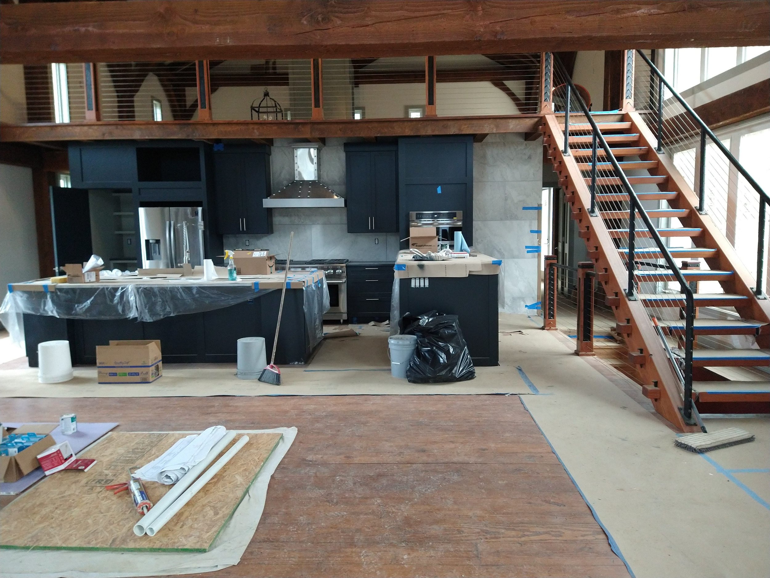 Kitchen under construction with a dark blue island, black cabinetry, stainless steel appliances, and a wooden staircase leading to a second-floor loft area.