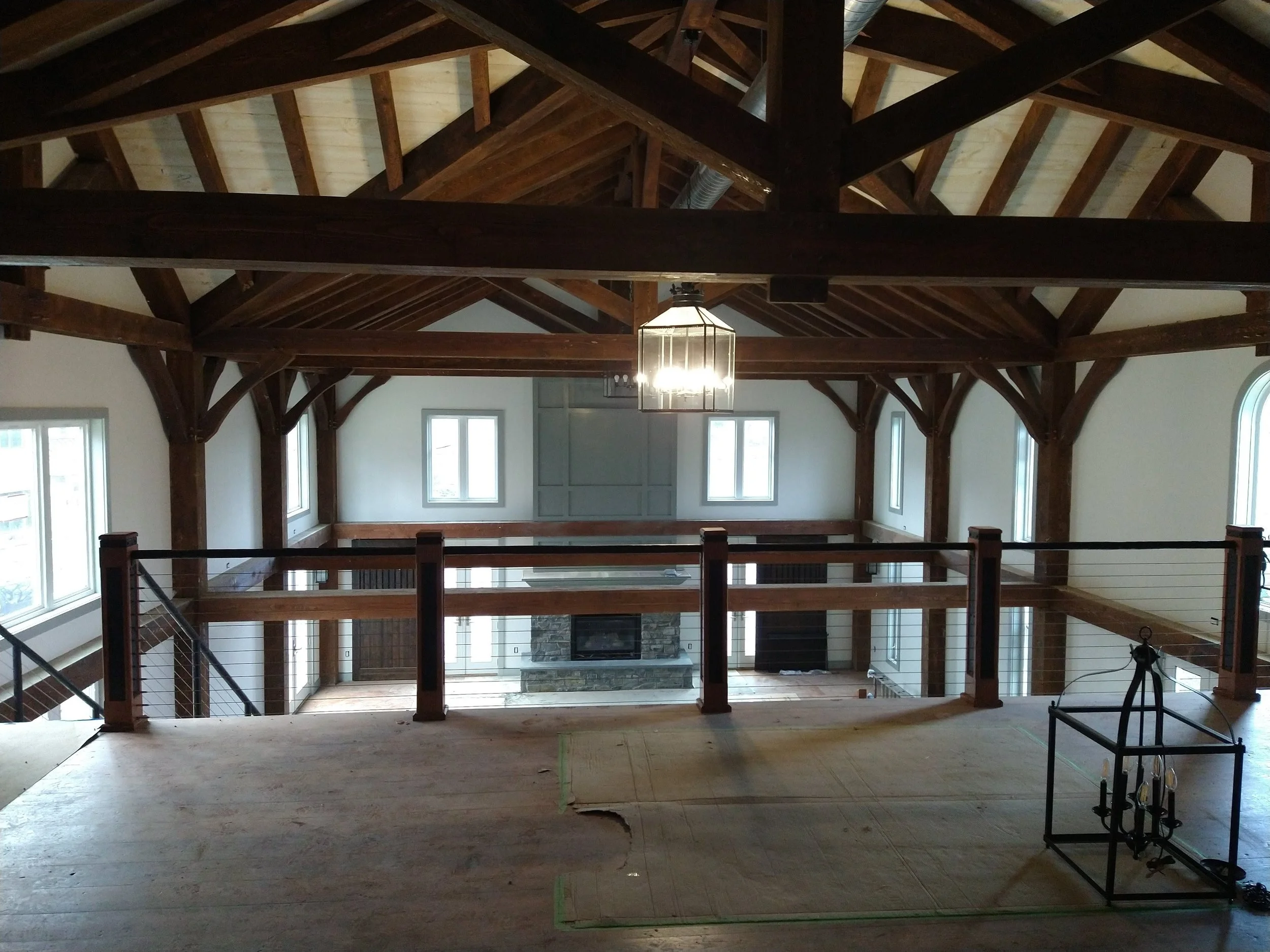 Interior of a large, open living room with a high vaulted ceiling supported by dark wooden beams, multiple windows on white walls, a chandelier hanging from the ceiling, and a fireplace with a stone surround.