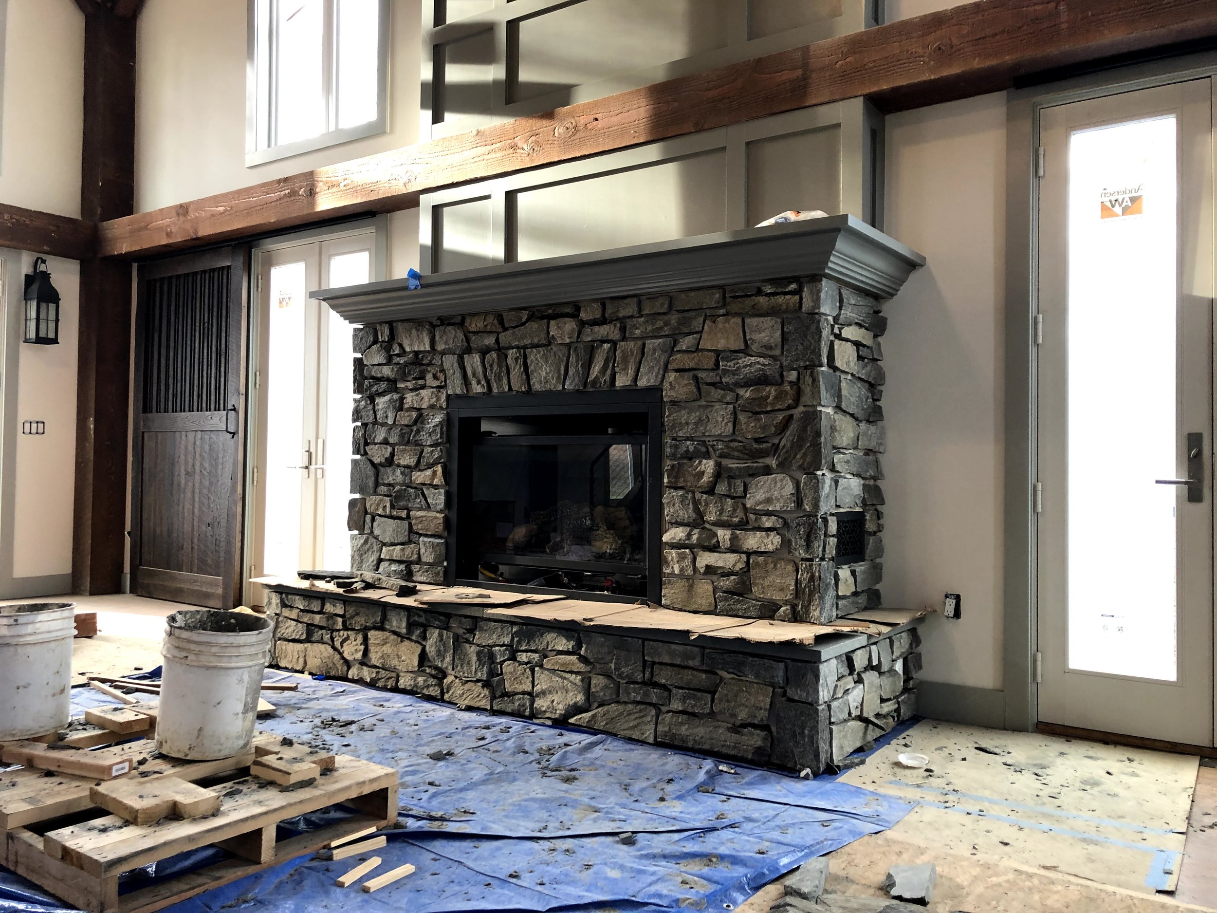Living room with a stone fireplace under construction, building materials and tools on the floor, and a door with a window to the outside.