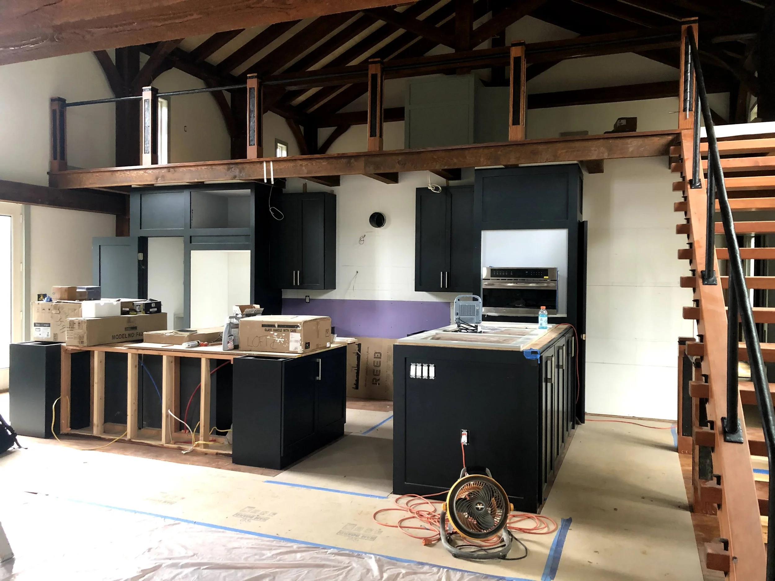 Interior of a house under renovation with a kitchen area, black cabinets, and a loft with a wooden staircase.