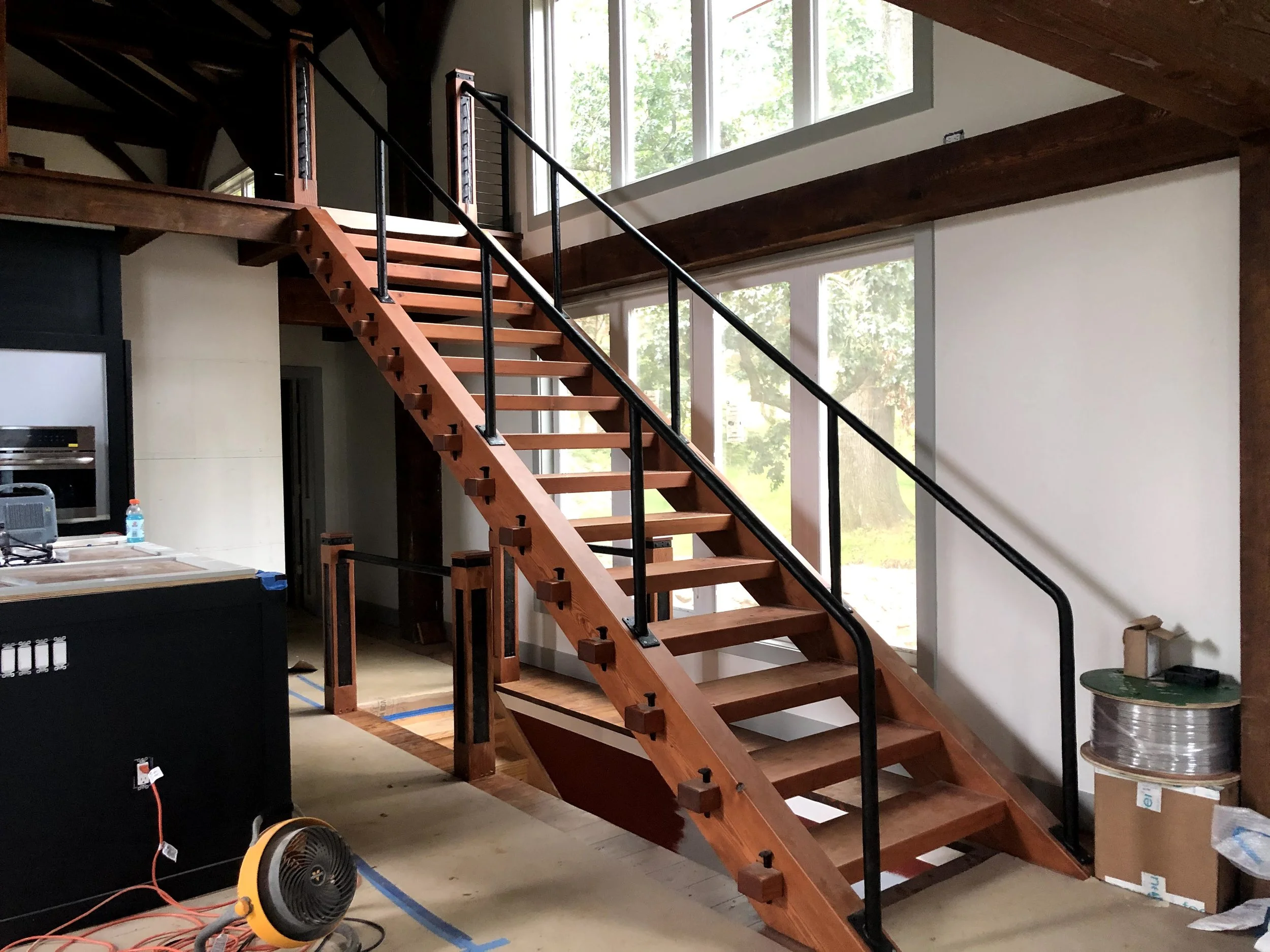 Interior view of a house under construction showing a custom open riser wooden staircase with black metal railings, and large windows letting in natural light.