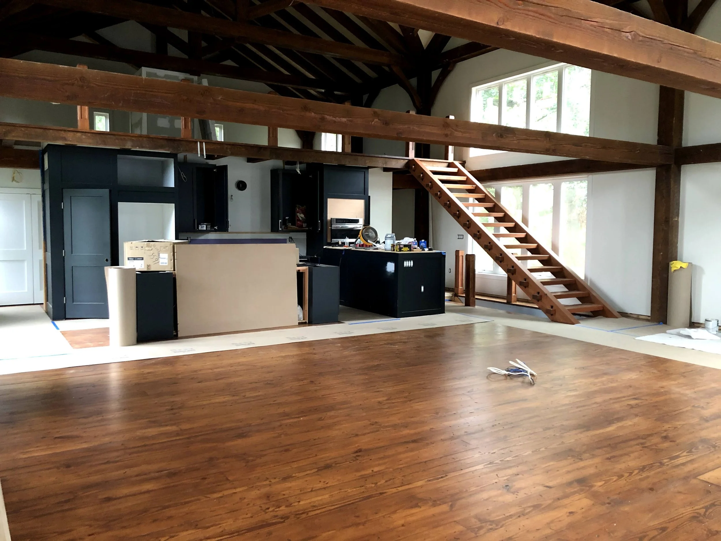 Interior view of a spacious room with high wood-beamed timber frame ceiling, kitchen area under construction, and large windows allowing natural light.