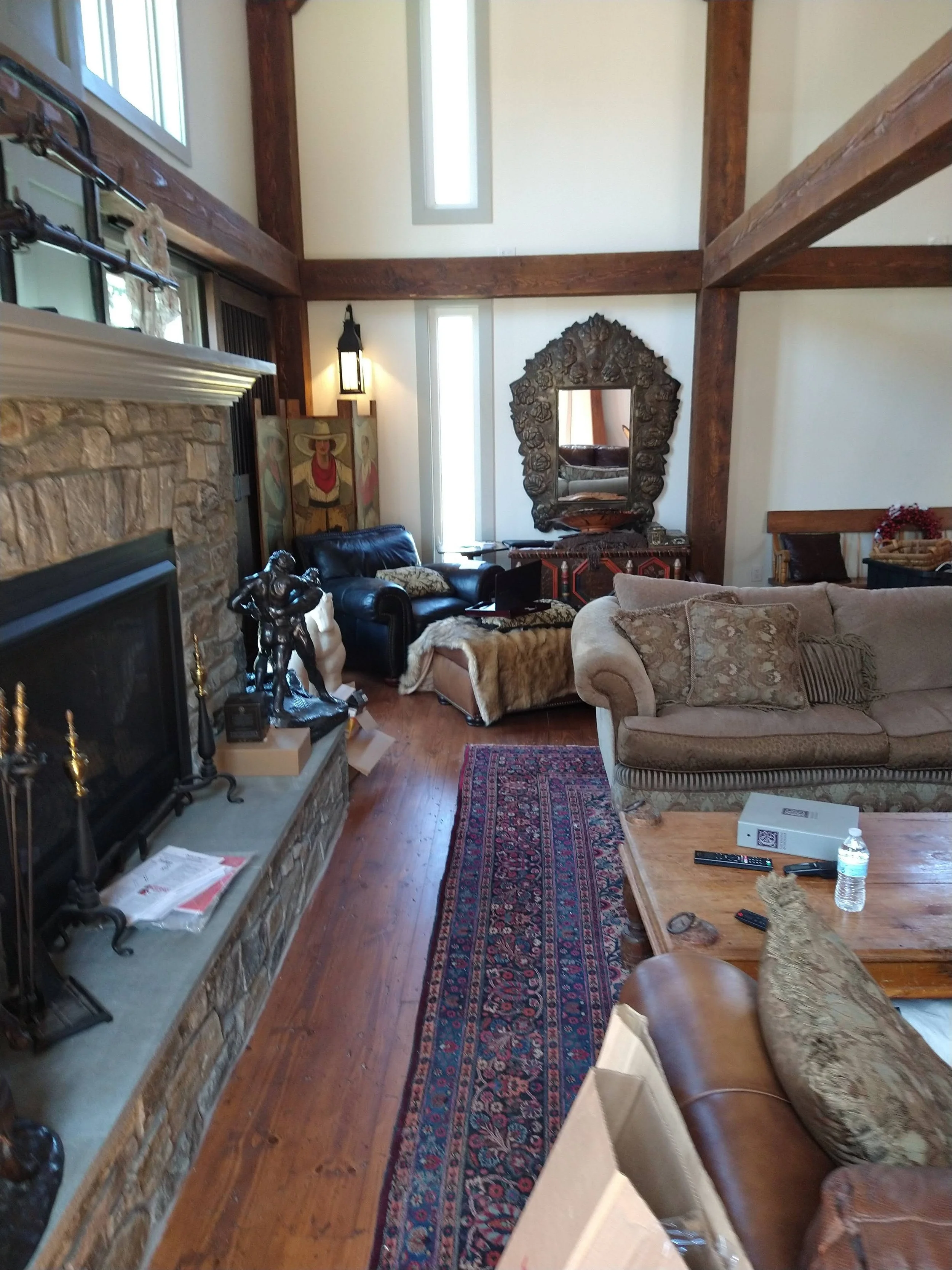 Living room with stone fireplace, large mirror, beige couch, black armchair, and wooden coffee table with water bottle and remote controls, decorated with art, pillows, and a patterned rug.