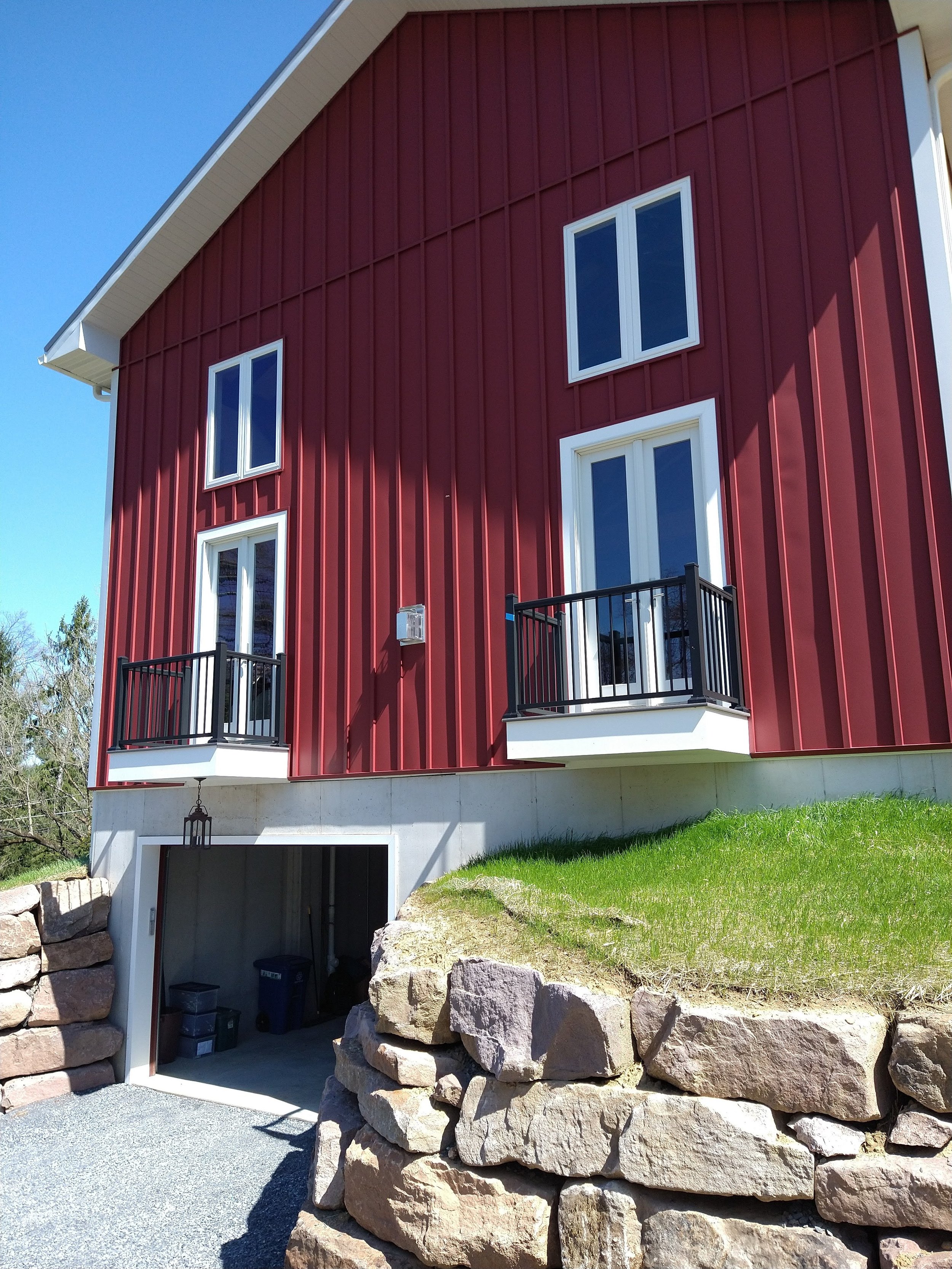 Red multi-story house with white trim, two small balconies with black railings, large windows, and an open garage at ground level, set on a grassy hill with stone retaining walls.