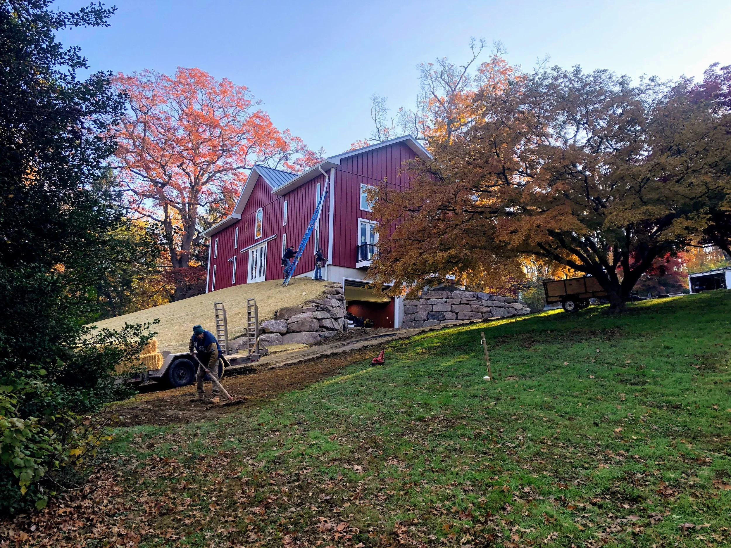 Workers are working on landscaping or construction on a sloped yard in front of a red house with white trim, surrounded by trees with fall foliage.
