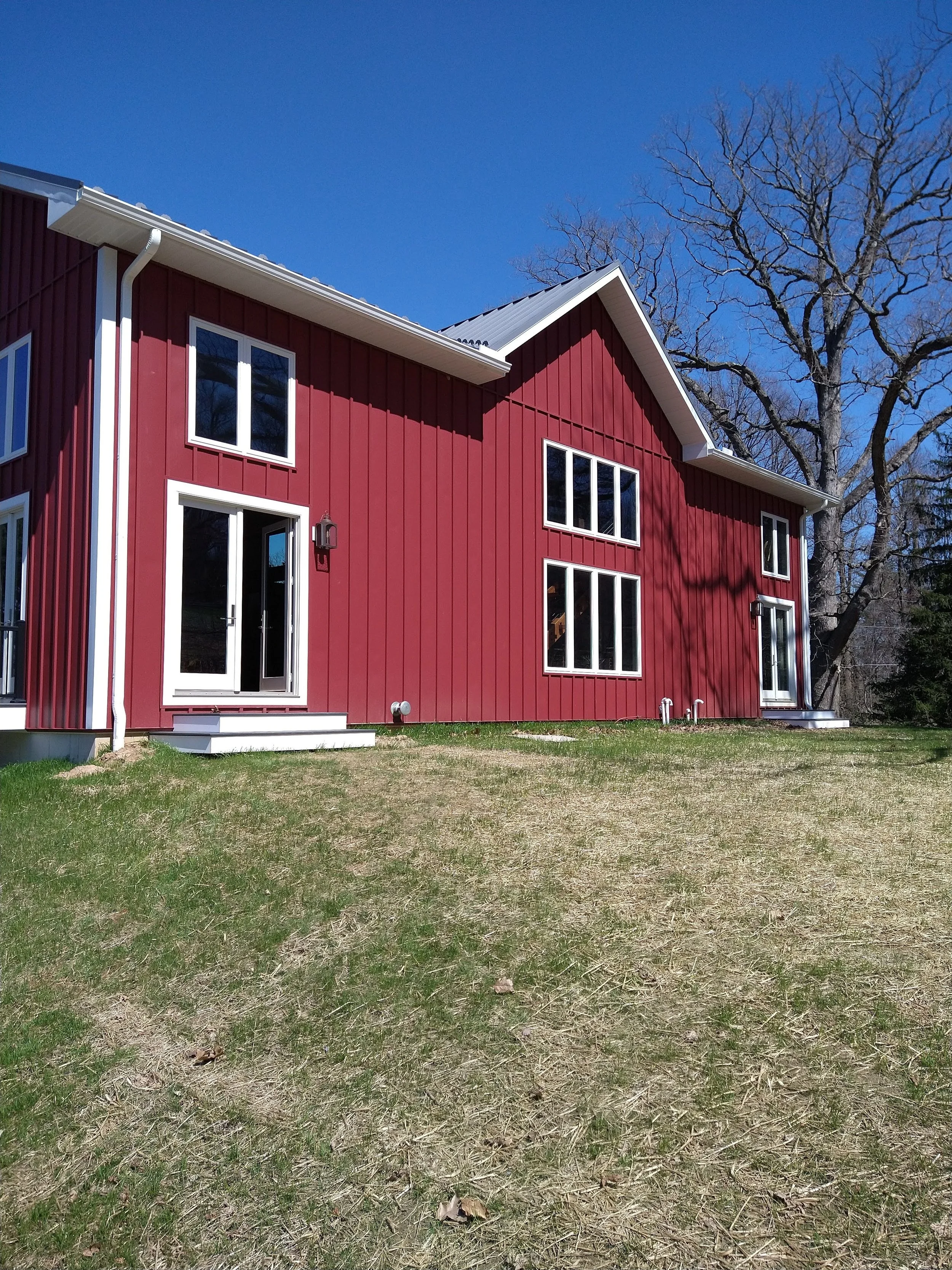 A two-story red house with white trim, multiple windows, and a small patio door, surrounded by a grassy yard and large leafless tree under a clear blue sky.