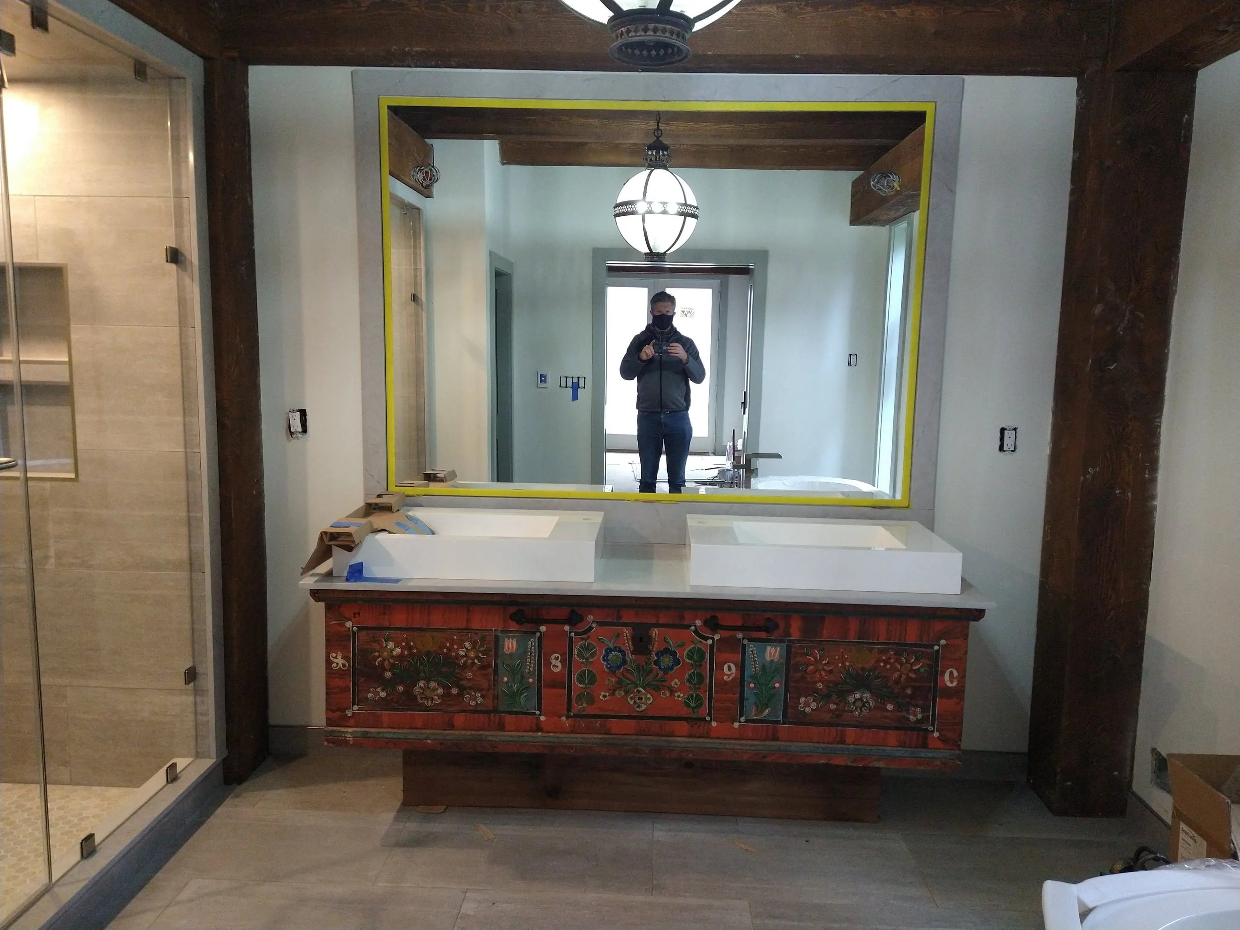 A bathroom vanity with two white sinks beneath a large rectangular mirror, with construction materials on the cabinet, in a room with a shower on the left and wood beams overhead.