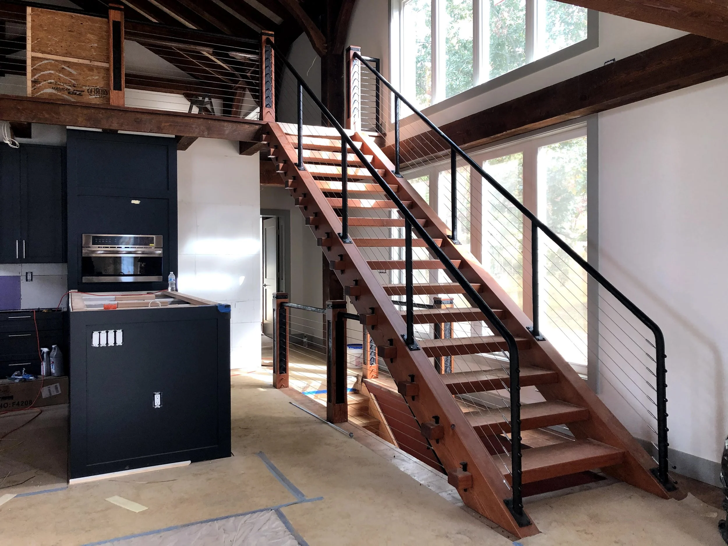 Interior view of a house under construction featuring wooden staircase with black metal railings leading to an upper loft area, large windows letting in natural light, partially visible kitchen with black cabinetry and appliances, and construction ma