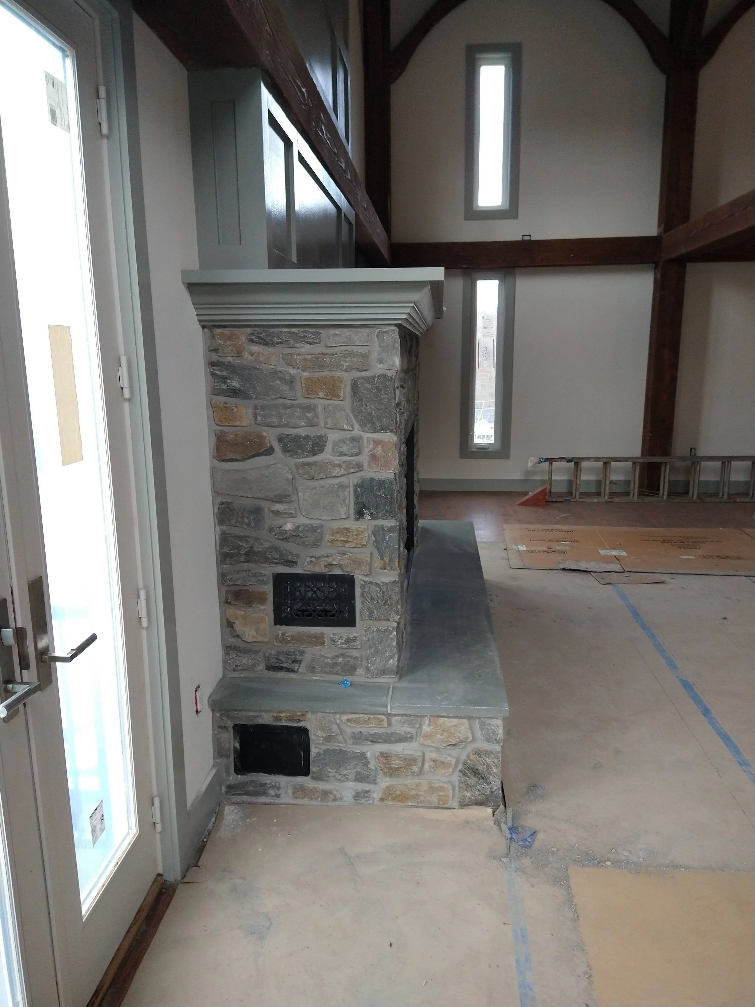 Interior of a house under construction, showing a stone fireplace with a wooden mantel, adjacent to a glass door and tall narrow windows, and framing on the floor and walls.