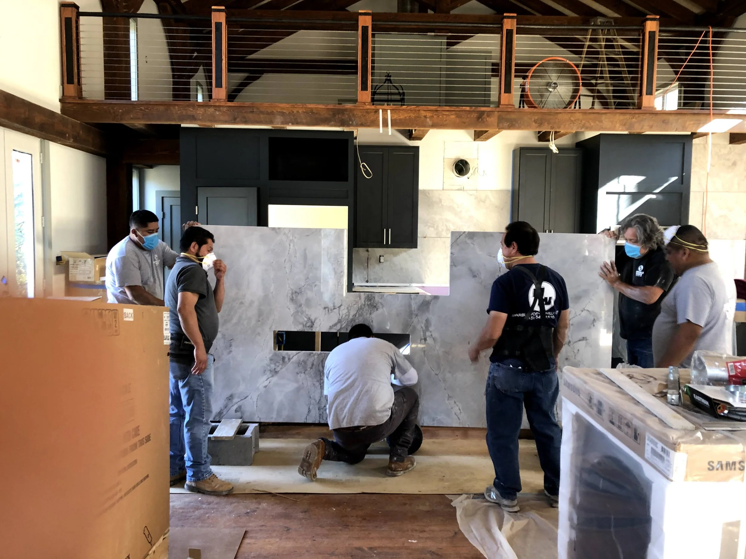 A group of seven workers wearing face masks and working together on installing a large marble or stone slab in a kitchen. Two workers are kneeling while others stand around the slab, which is positioned in front of black cabinetry and a kitchen openi
