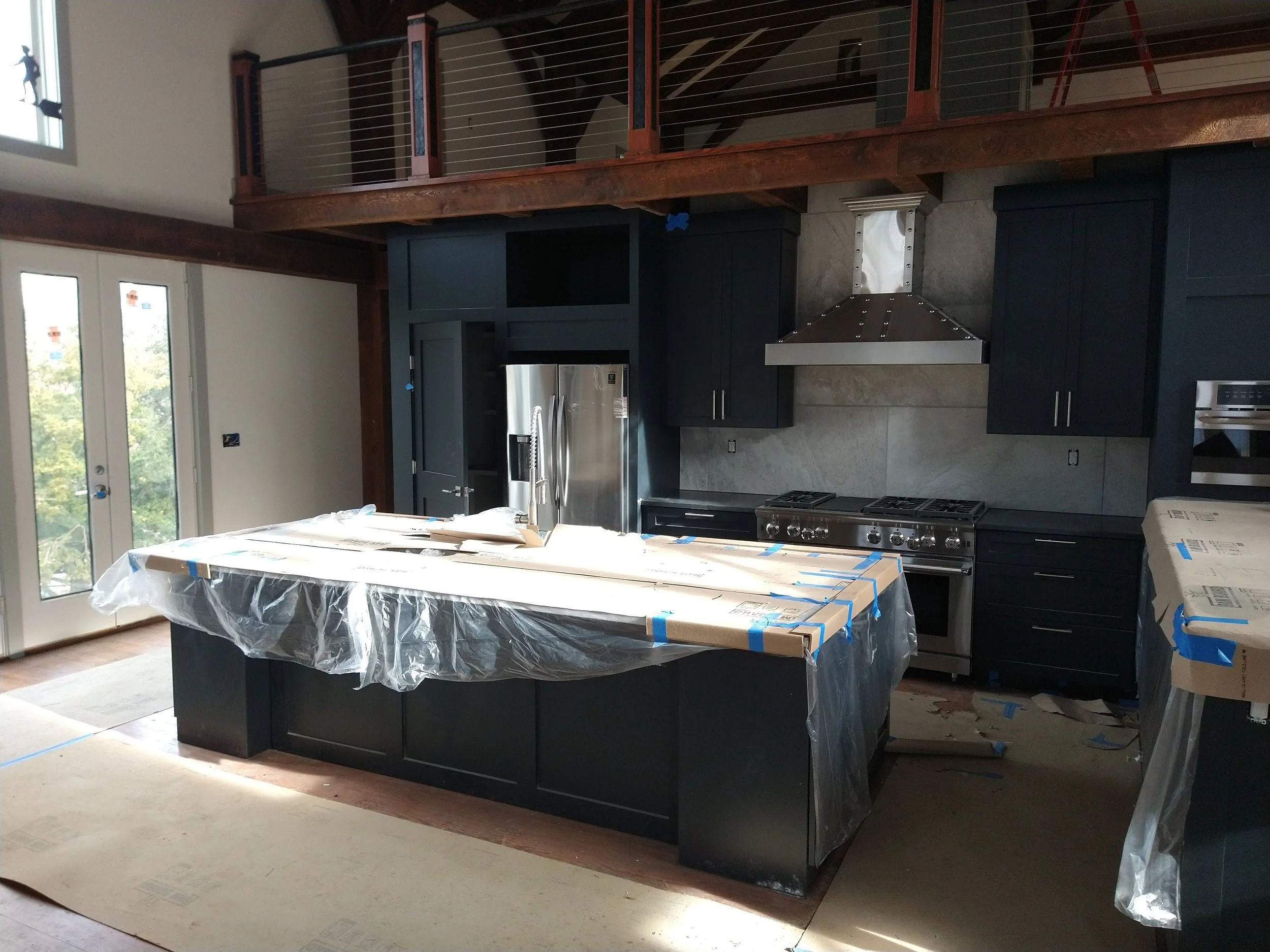 Kitchen under construction with black cabinets, stainless steel refrigerator, and a large island covered in plastic and cardboard.