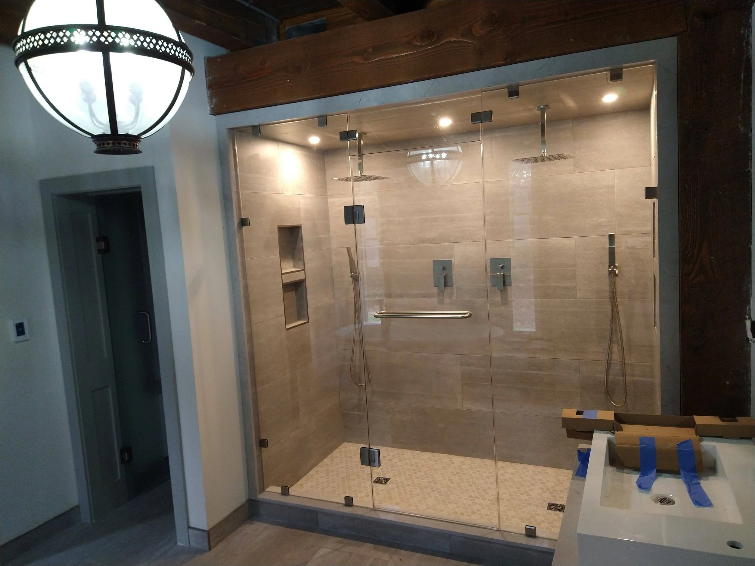 Unfinished walk-in shower with glass doors in a bathroom, featuring two overhead rain showerheads and handheld shower wands, beige tiles, and built-in niches on the wall.