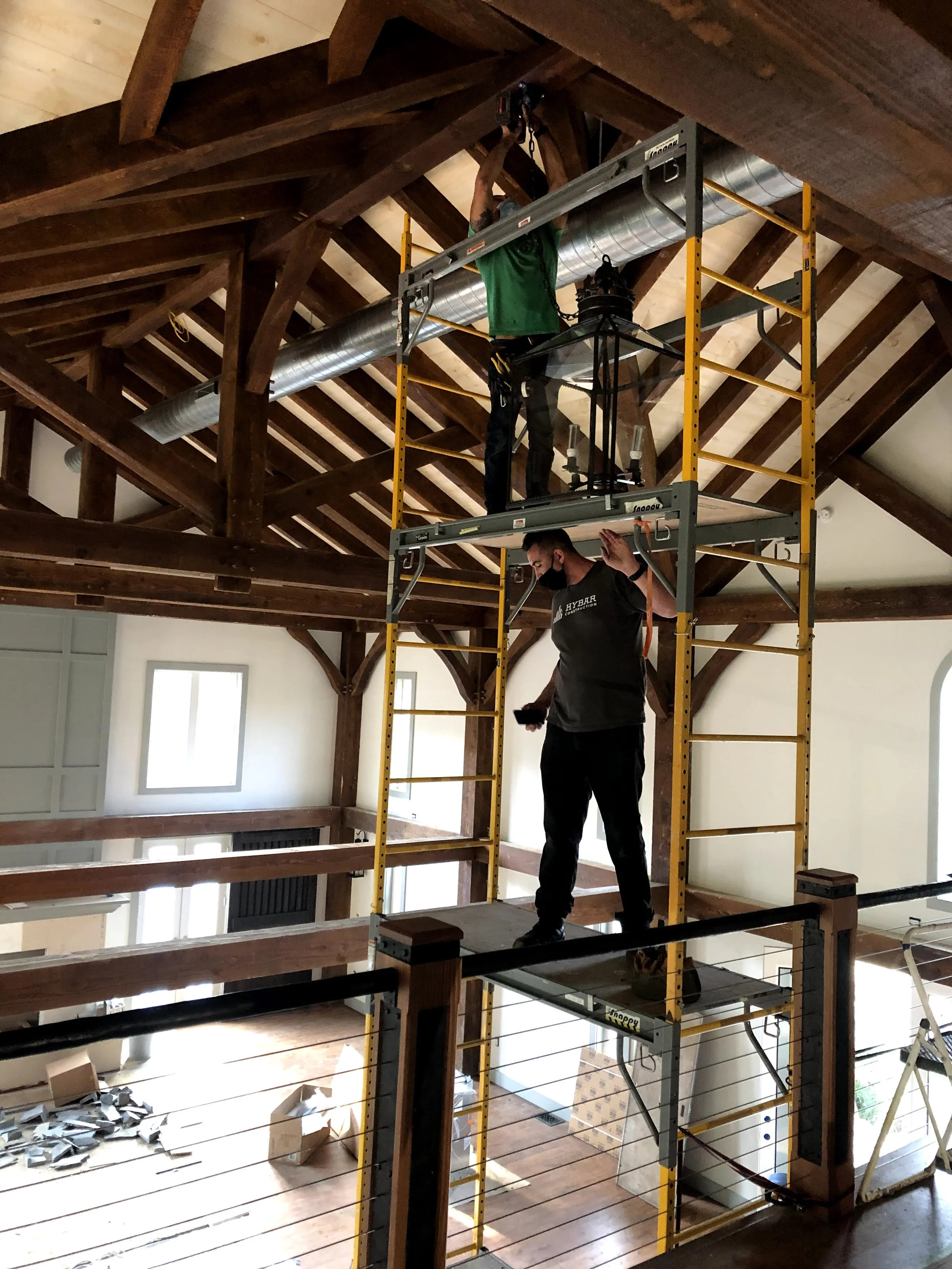Two workers installing ductwork and lighting on the ceiling of a wooden loft space. The space has large windows and a partially finished interior.
