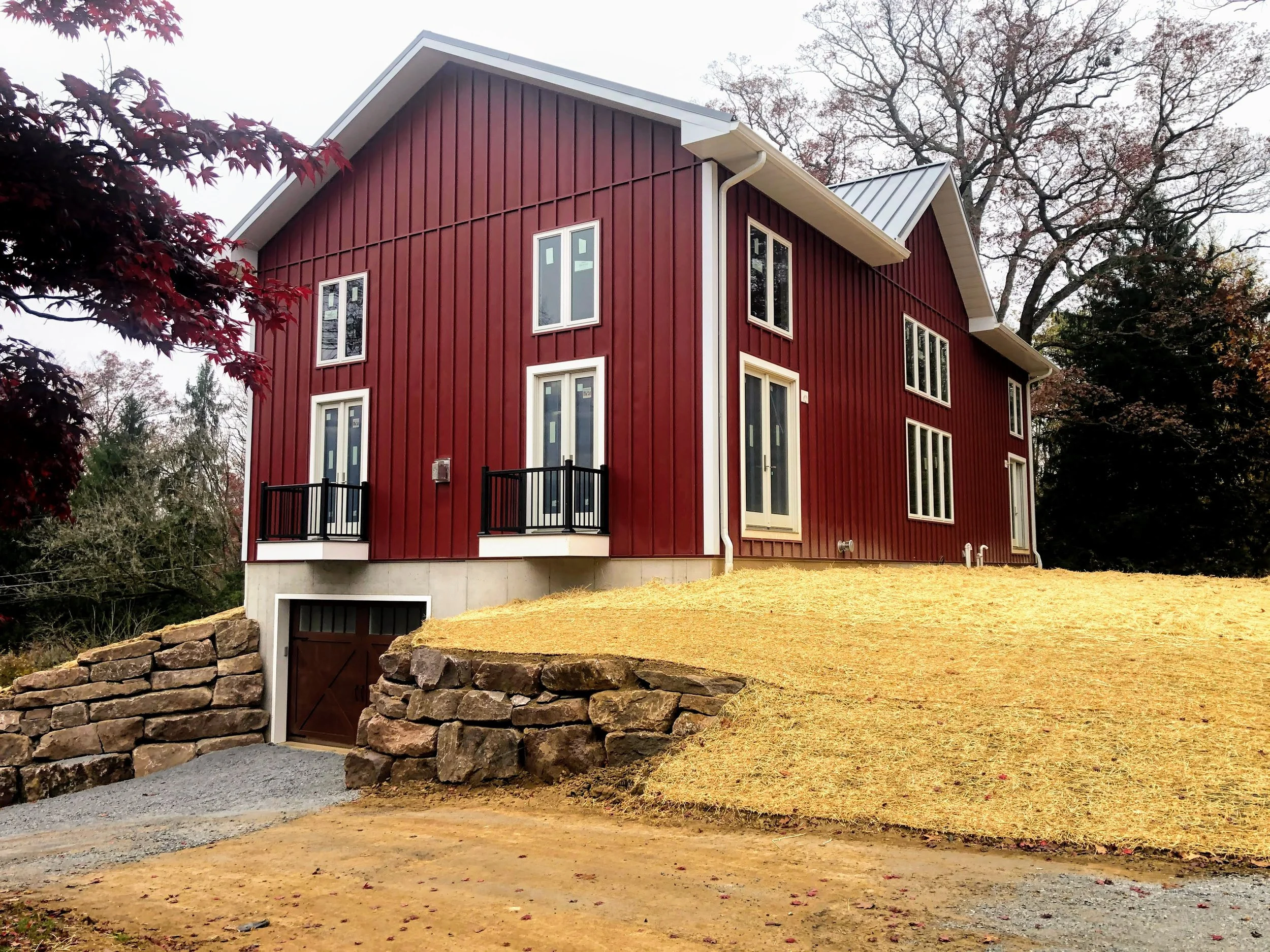 A red multi-story house with white trim, black balcony railings, and a garage with stone retaining walls leading up to a driveway, set on a grassy hill with trees in the background.