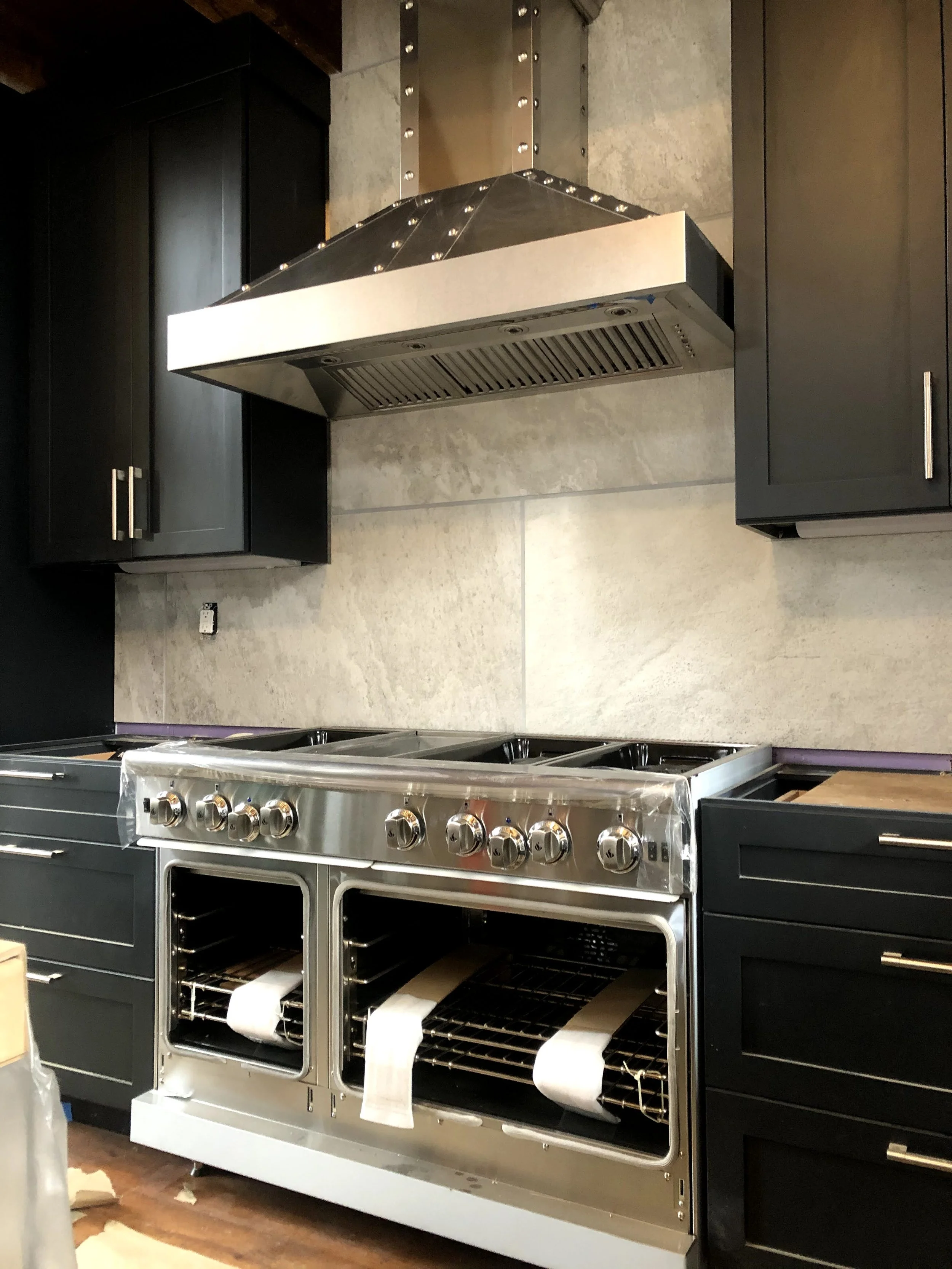 A modern kitchen with black cabinetry, a large stainless steel oven with multiple burners, a stainless steel range hood, and a beige stone backsplash.