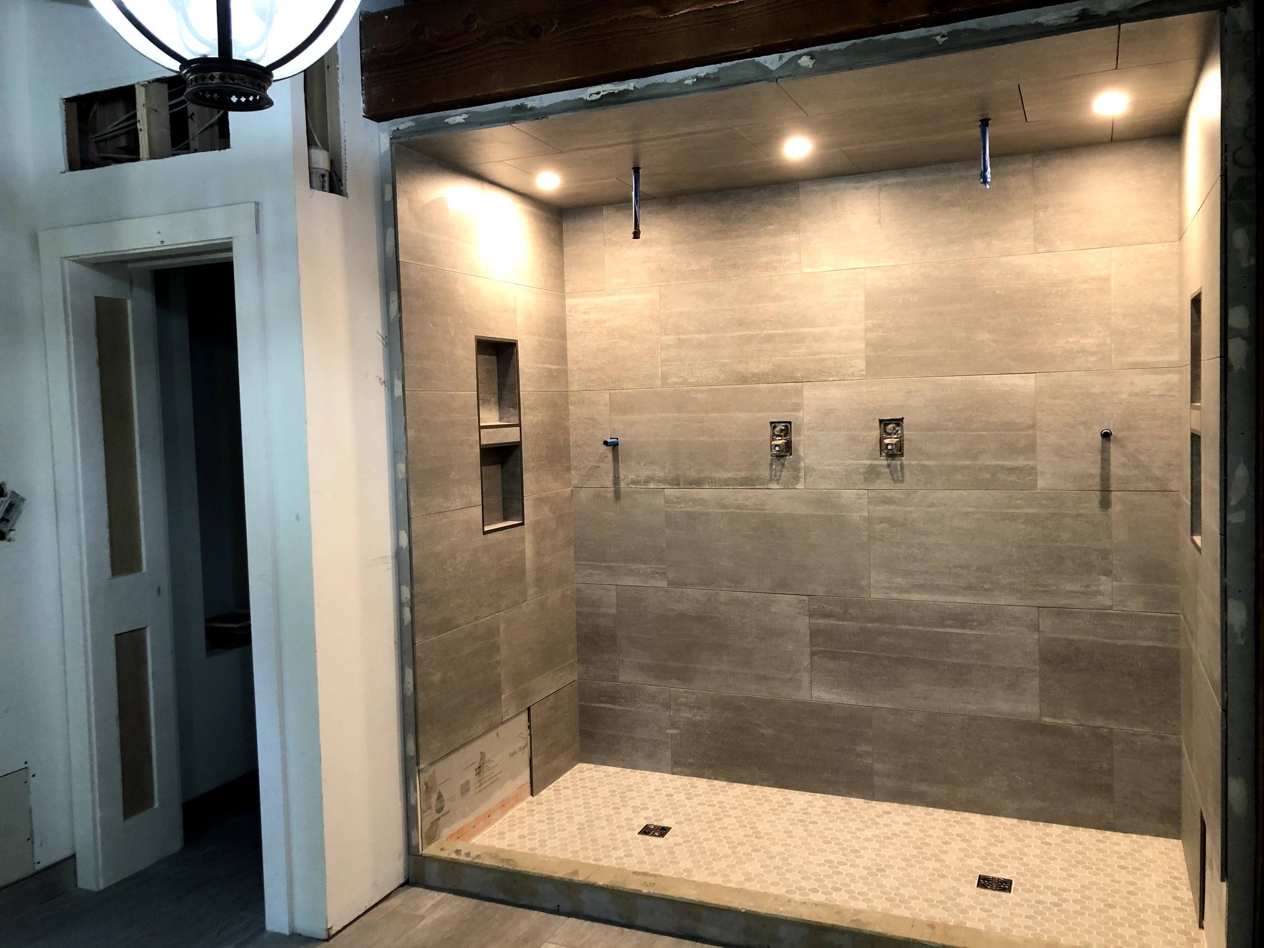 Interior of a partially finished bathroom shower with tiled walls, small recessed shelves, and lighting fixtures installed on the ceiling.
