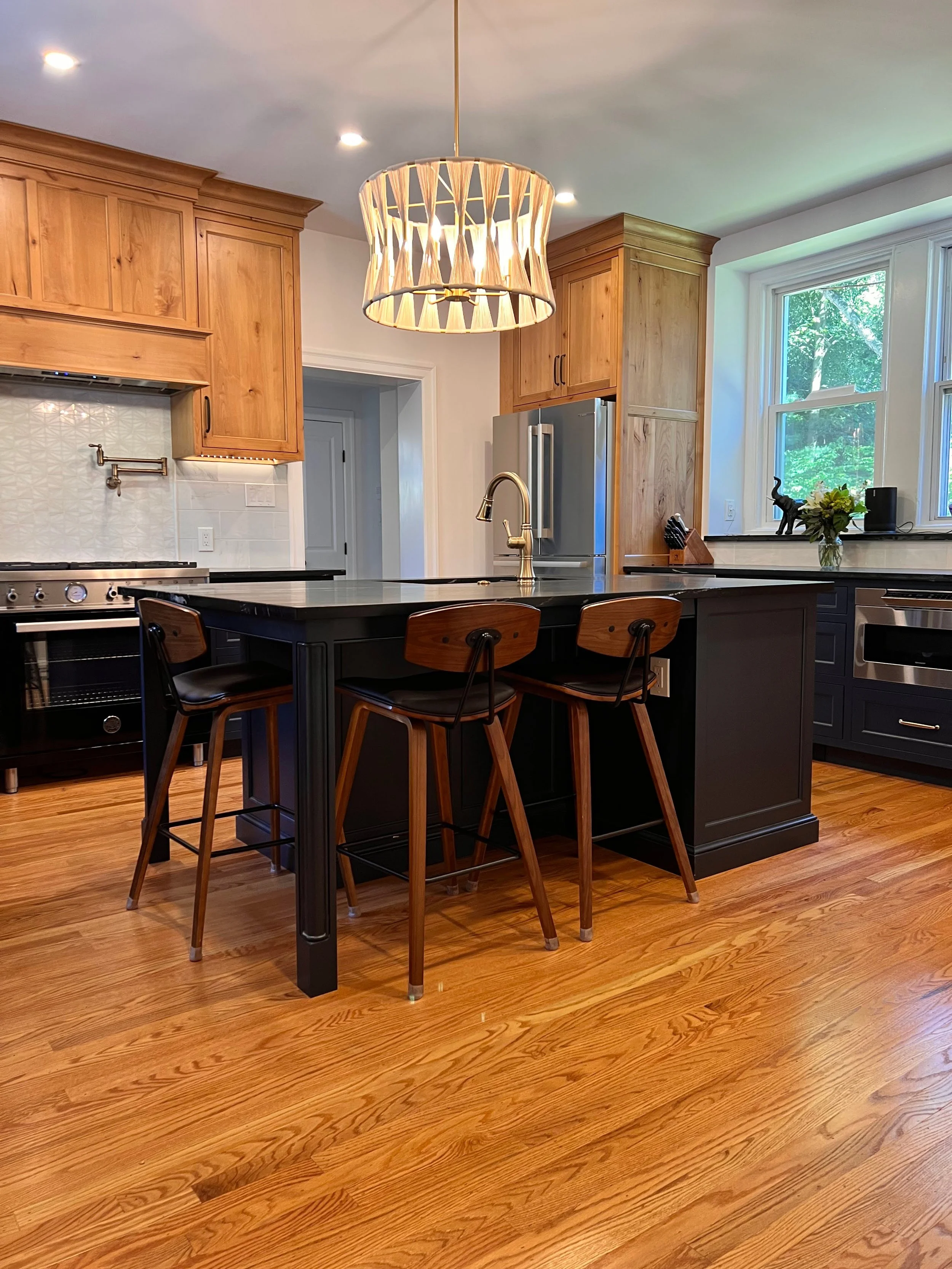 Modern kitchen with wooden cabinets, black island, two bar stools, pendant light, stainless steel appliances, hardwood floor, and large window.