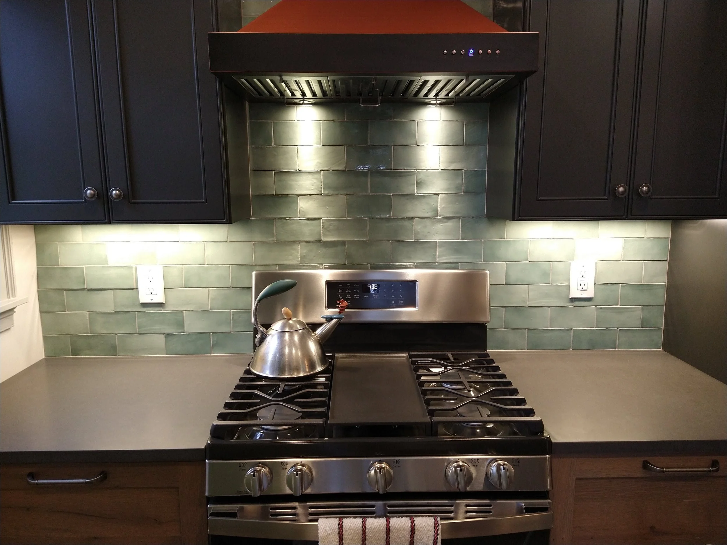 Modern kitchen with stainless steel gas stove, green tiled backsplash, kettle on stove, black cabinets, and overhead range hood with lighting.