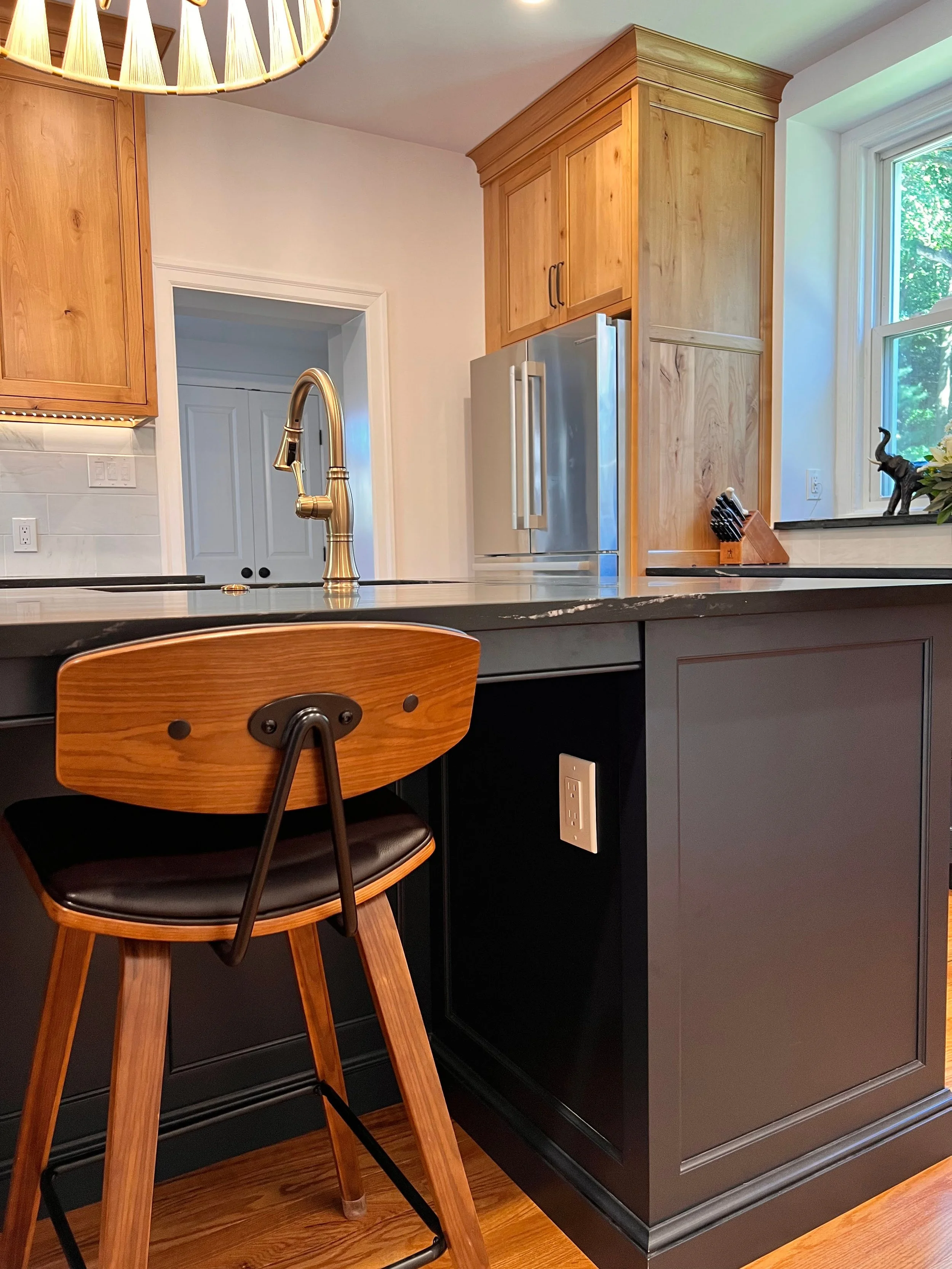 Modern kitchen with wooden cabinets, stainless steel refrigerator, marble countertop, and a brass faucet. A wooden barstool is at the island, and a decorative light fixture hangs from the ceiling.