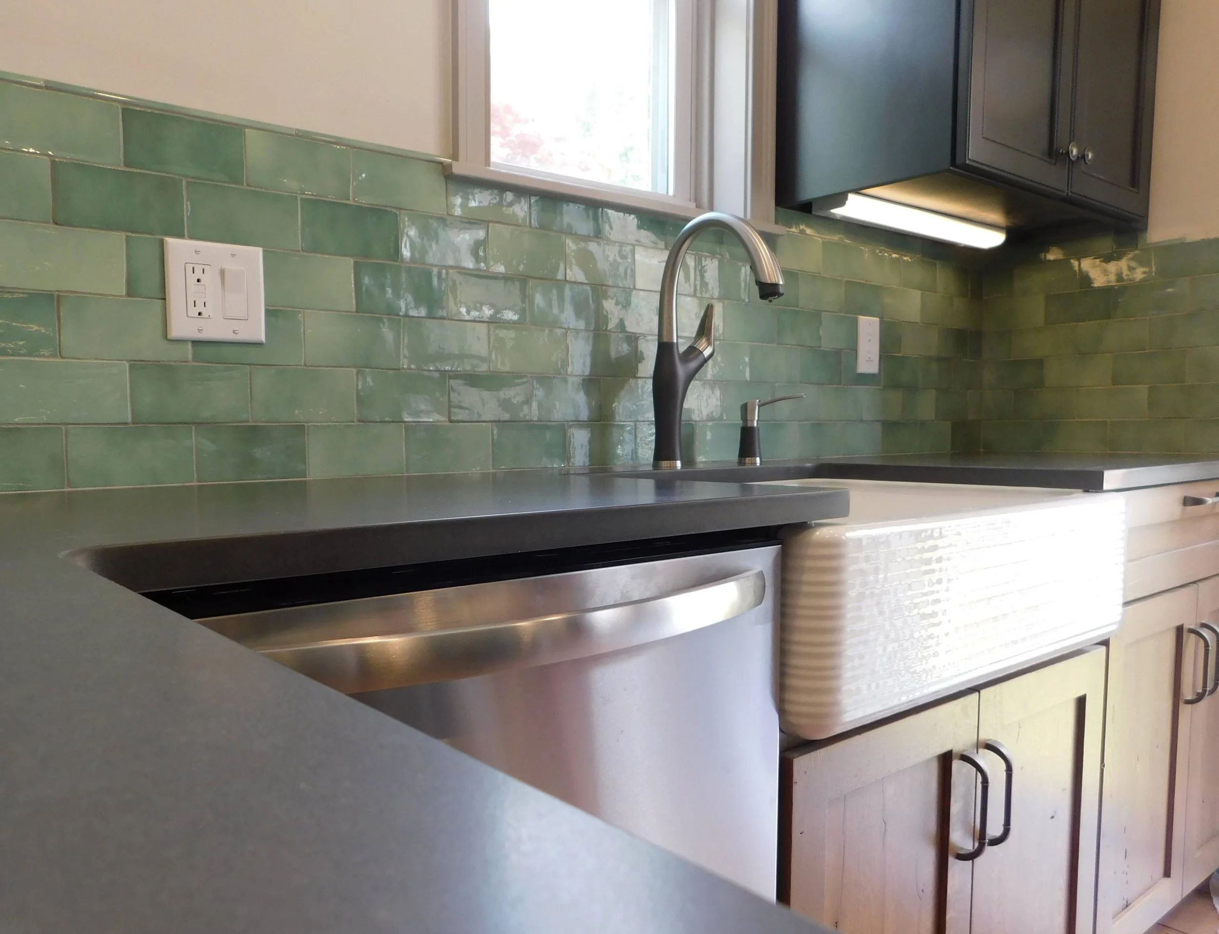 Modern kitchen interior with green tile backsplash, stainless steel faucet, farm-style sink, dark countertop, and wooden cabinets.