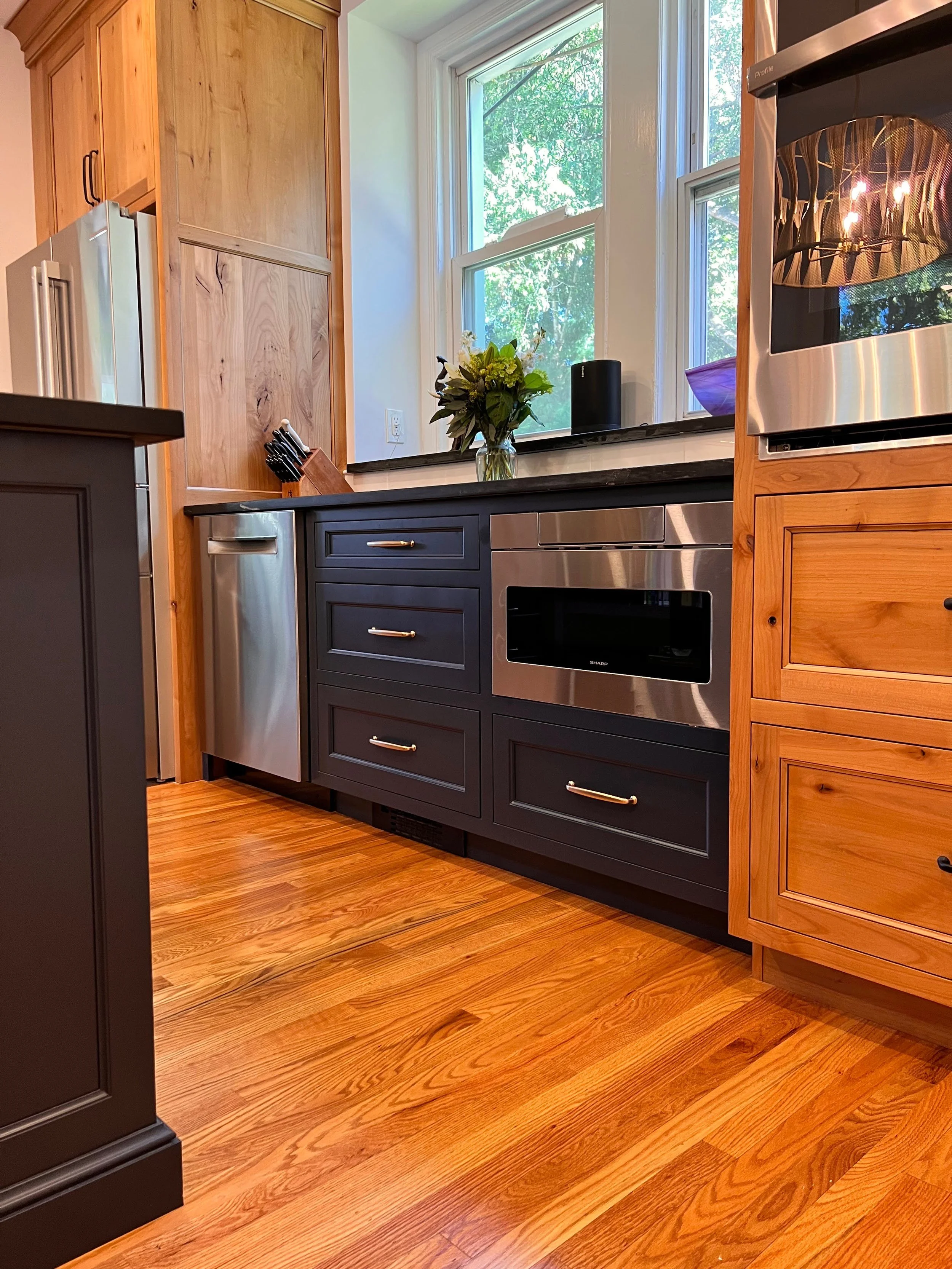 Modern kitchen with wooden cabinets, stainless steel appliances, and a wooden floor. A vase with flowers on the countertop and a knife block next to a window are visible.