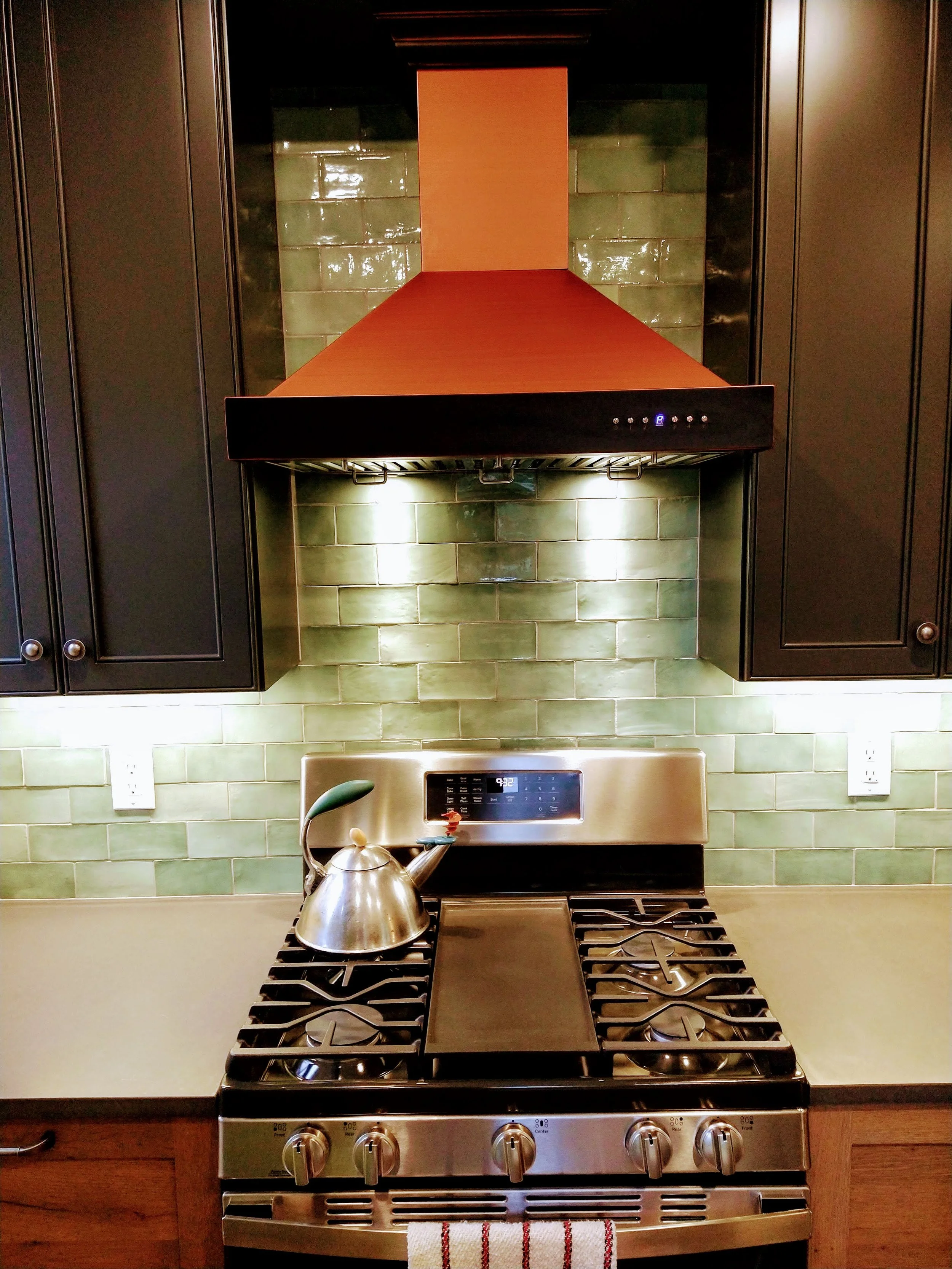 Stainless steel gas stove, black grill and control knobs at the front, green tiled backsplash, black upper vent hood, and dark cabinets on either side.