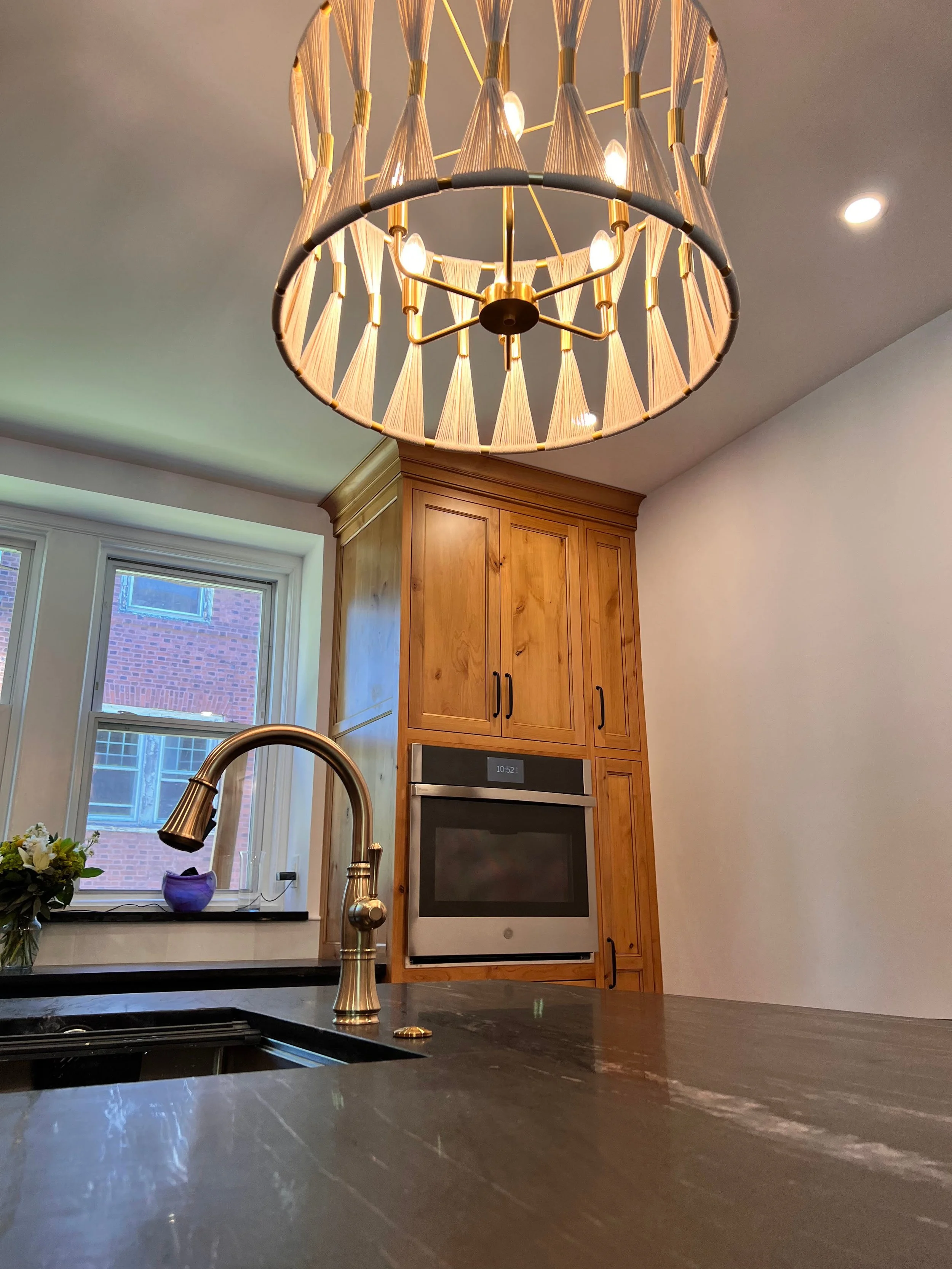 Modern kitchen with brass faucet and large wooden cabinets, overhead light fixture, black countertop, built-in oven, and window.