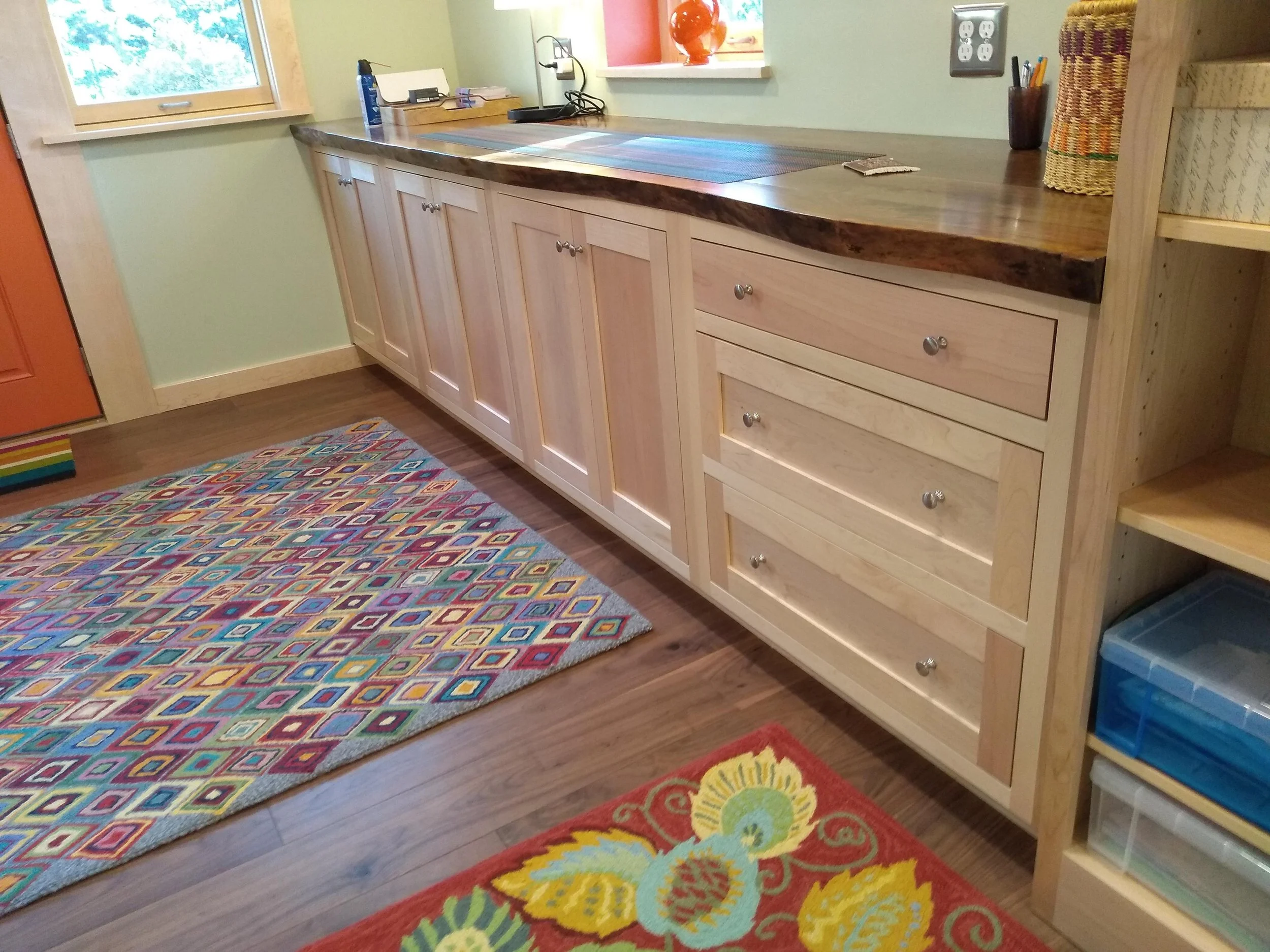 Wooden cabinets with a live edge countertop, colorful patterned rugs, and storage shelves with containers in a well-lit room.