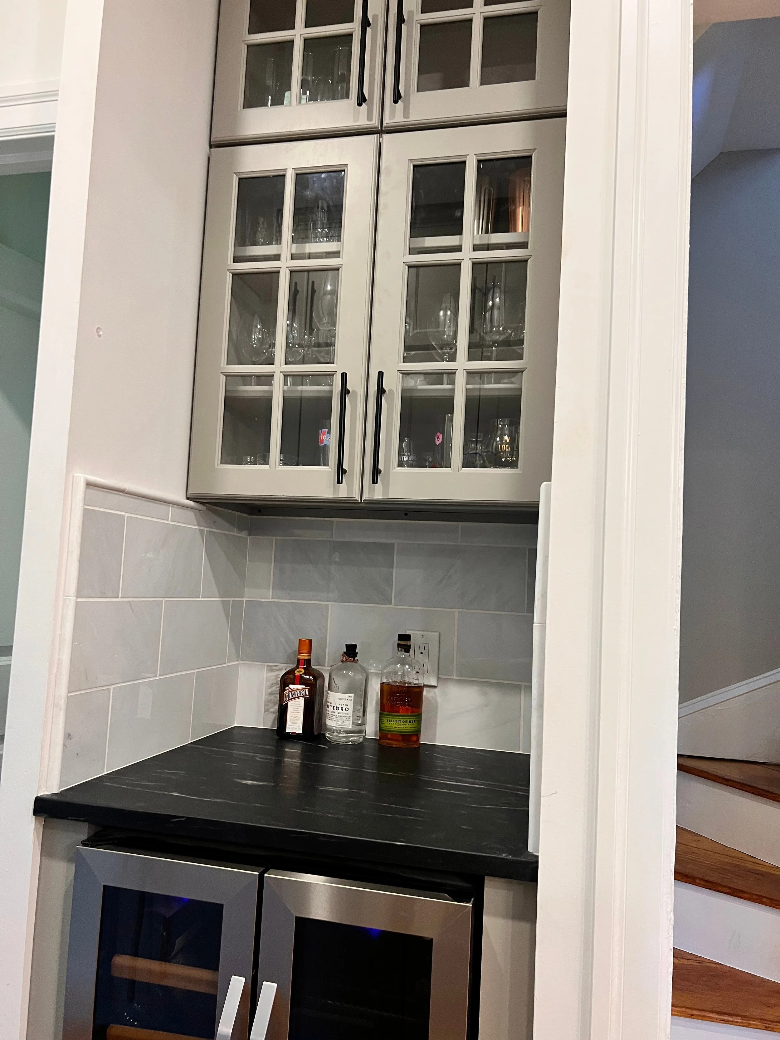 Kitchen bar area with a black countertop, bottles of alcohol, glass cabinets above, and a stainless steel wine cooler below.