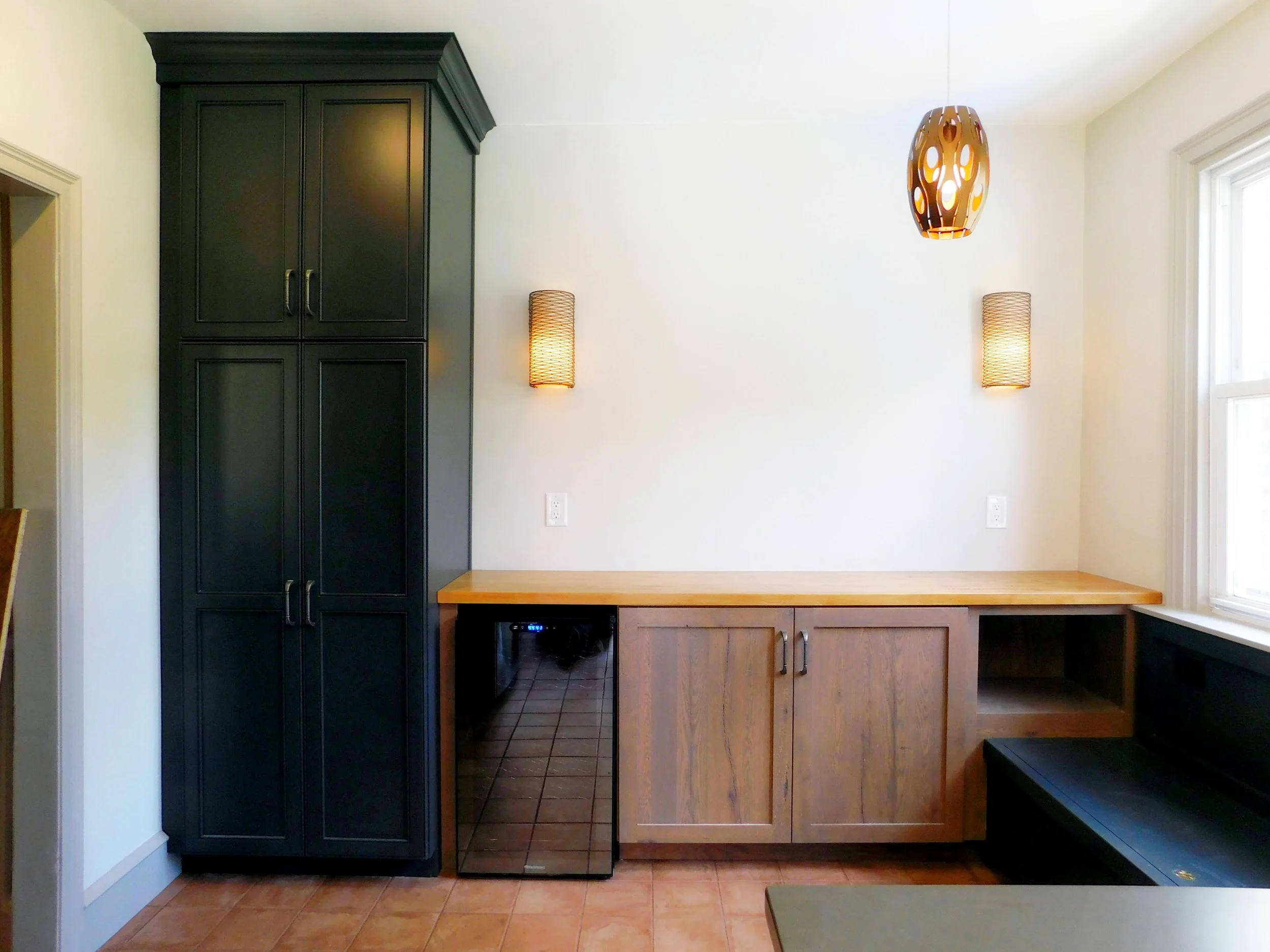 Modern kitchen interior with black tall cabinet, wooden countertop, built-in lower cabinets, and wall lights. Pendant light is hanging from the ceiling.
