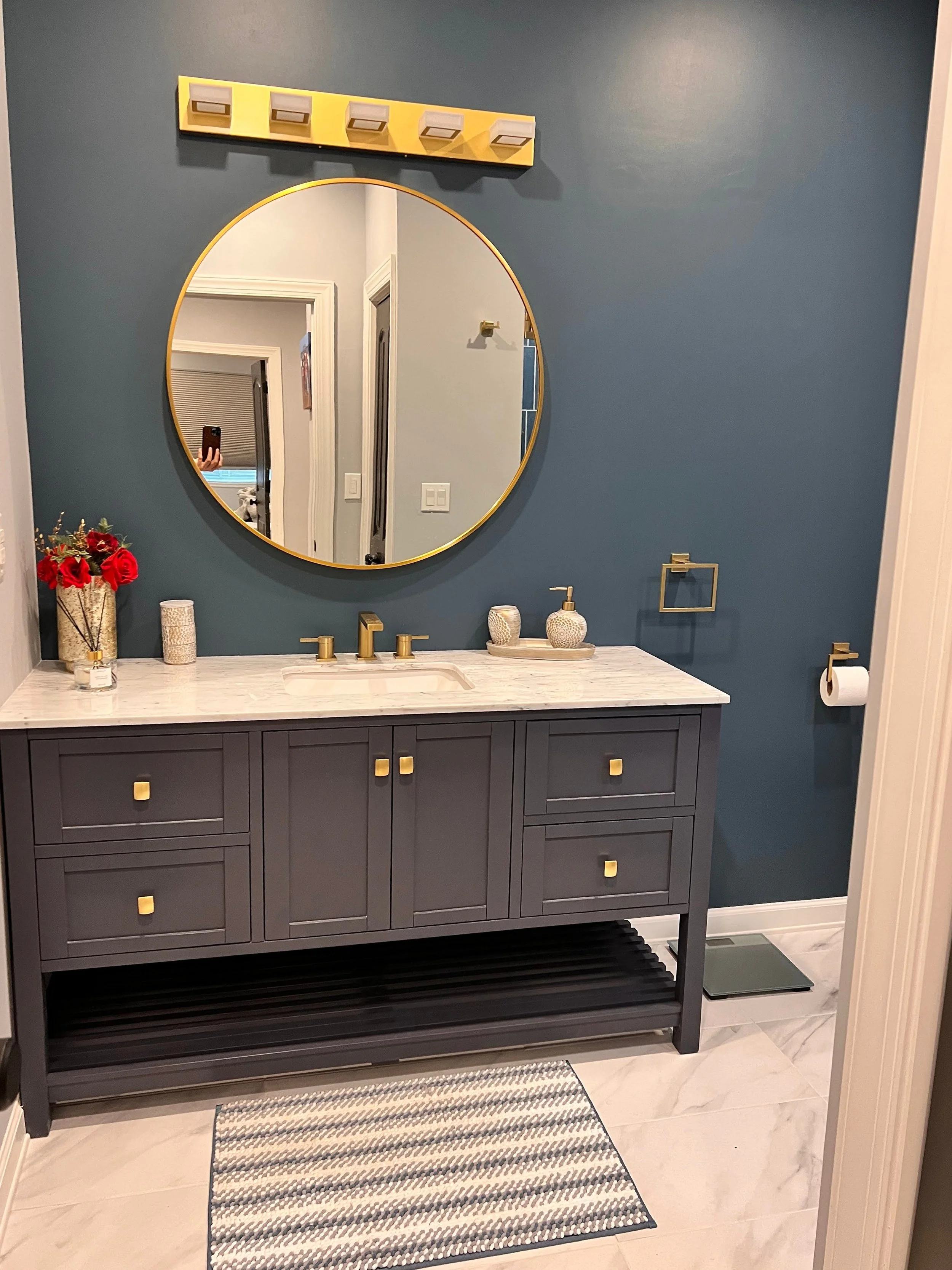 Modern bathroom with blue wall, gold accents, a dark gray vanity with a marble top, a round mirror, a vase with red flowers, decorative containers, and a striped rug on a light tile floor.