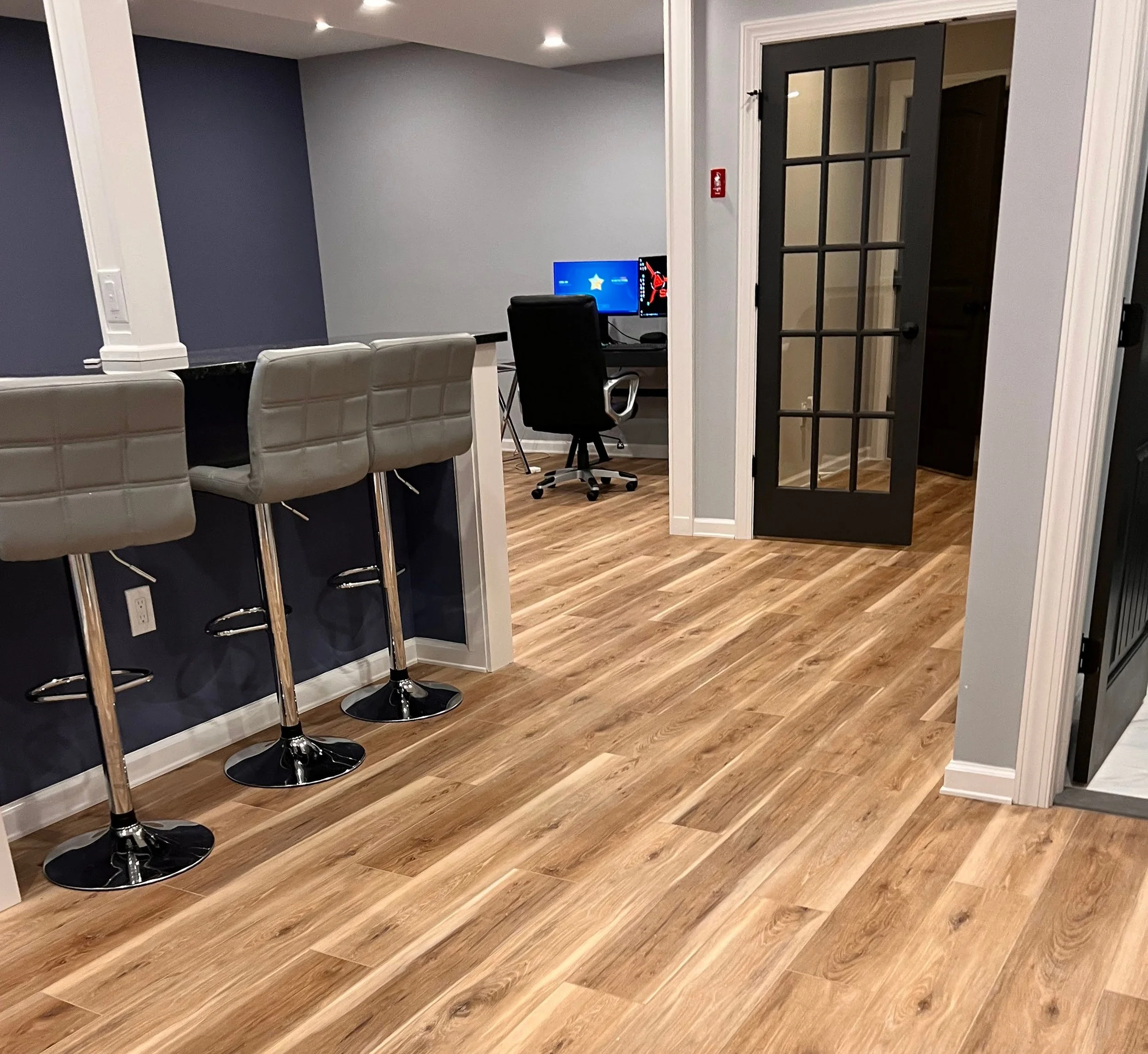 Home office with a small bar counter, three bar stools, a desk with a computer monitor, and a French door leading to another room, all with wood flooring and neutral wall colors.