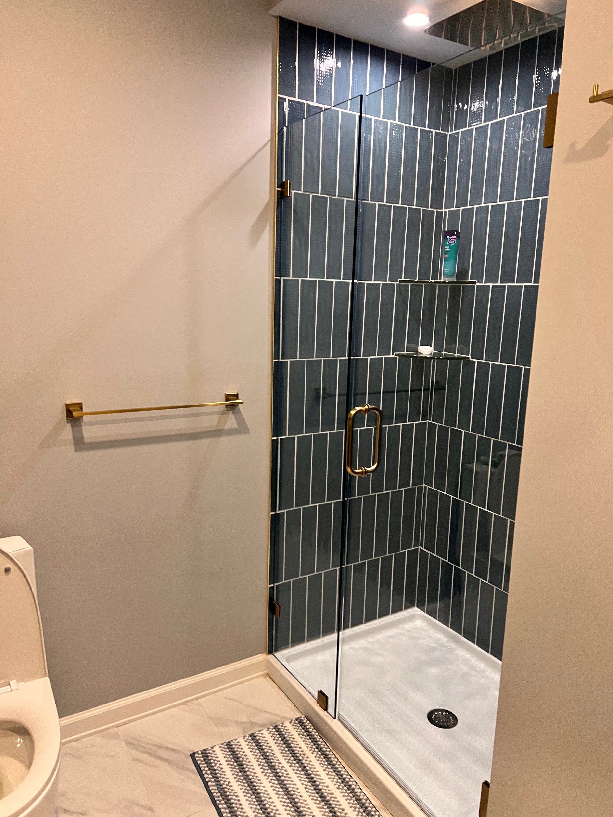 Bathroom with a glass-enclosed shower featuring dark blue tile walls, a gold handle on the door, a small shelf with soap and shampoo, and a floor drain. To the left, part of a toilet and a beige towel rack on the wall. A striped rug on the marble flo