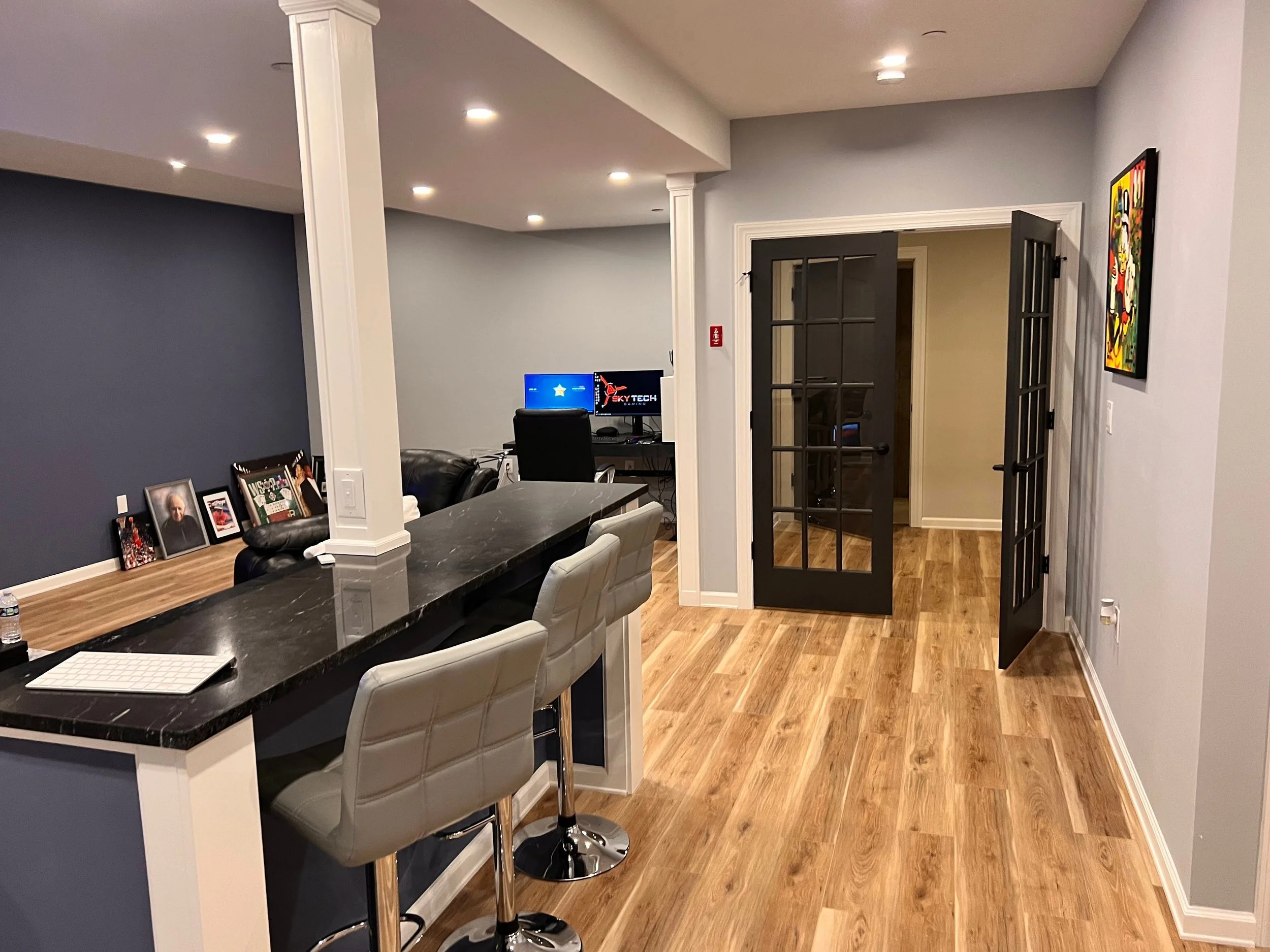 Interior view of a modern living space with a kitchen counter, bar stools, a small workspace with computers, and a room with glass-paneled black doors. The room features hardwood floors, colorful wall art, and a dark blue accent wall.