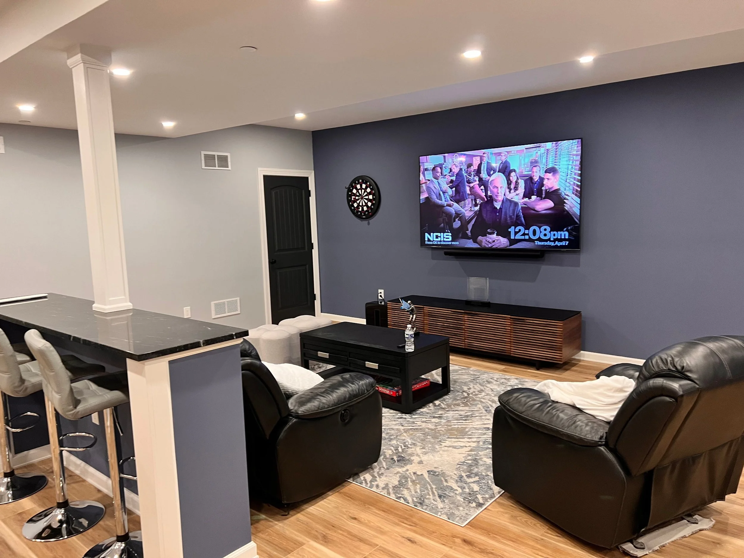 Living room with a large wall-mounted TV displaying a show and the time, a dartboard on the wall, a black entertainment console, leather recliners, a patterned area rug, a black coffee table, and a bar counter with barstools.