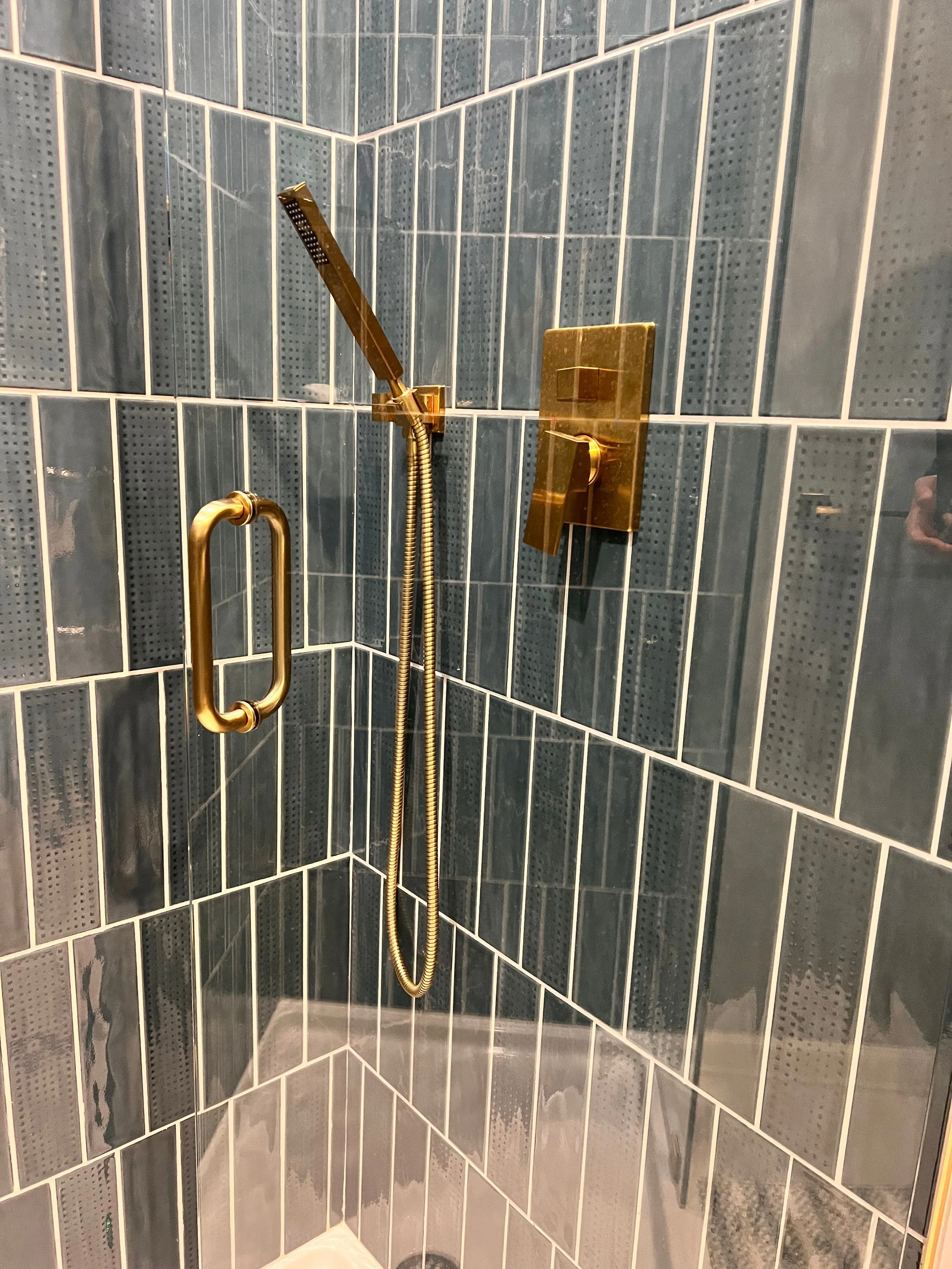 Shower with dark blue tiles, brass handle, and gold fixtures including a handheld showerhead and a wall-mounted control.