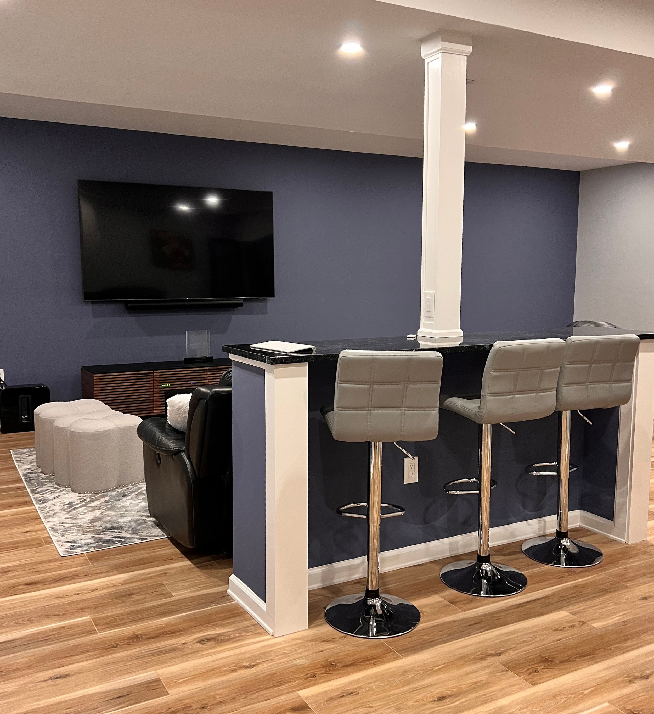 Modern living room with a mounted flat-screen TV, a dark wood media console, a black recliner, a gray sofa, a coordinating area rug, and a white kitchen counter with three gray barstools.