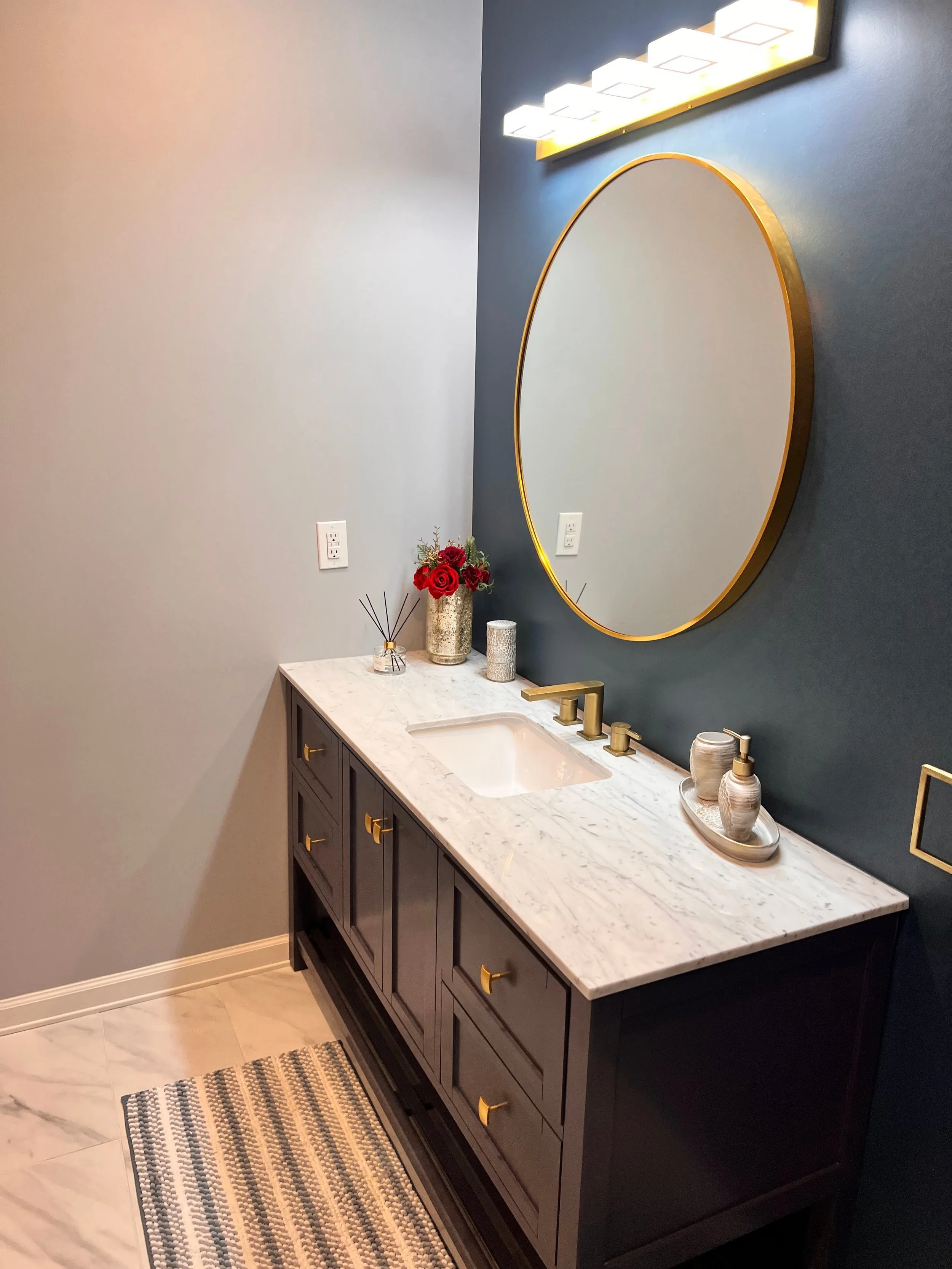 Bathroom vanity with a dark cabinet, white marble countertop, oval mirror with gold frame, flower vase with red roses, soap dispensers, and decorative items, with a modern light fixture above.