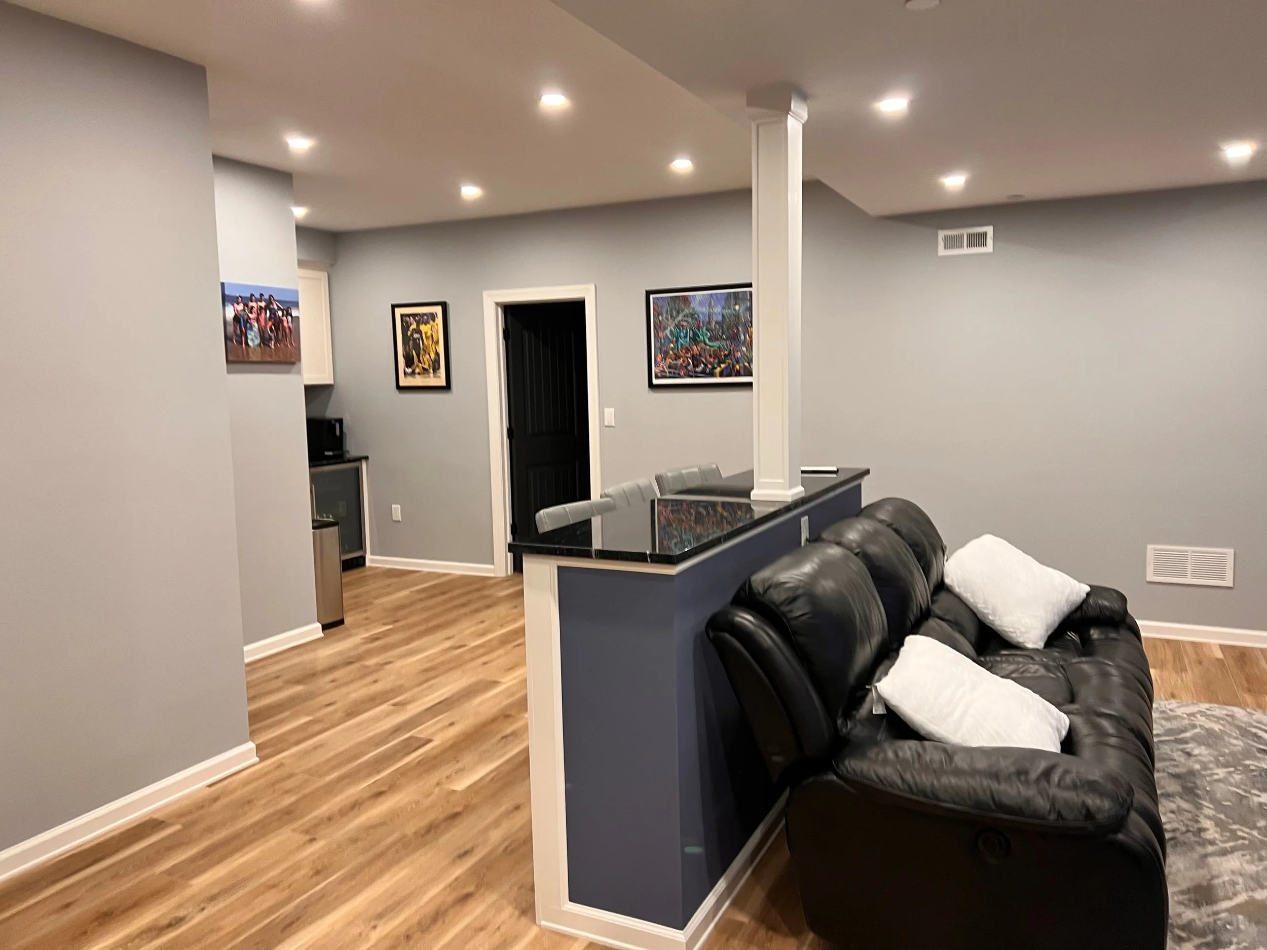 Living room with black leather couch, white pillows, wooden floors, grey walls, framed artwork, doorway, small kitchenette in background, recessed ceiling lights.