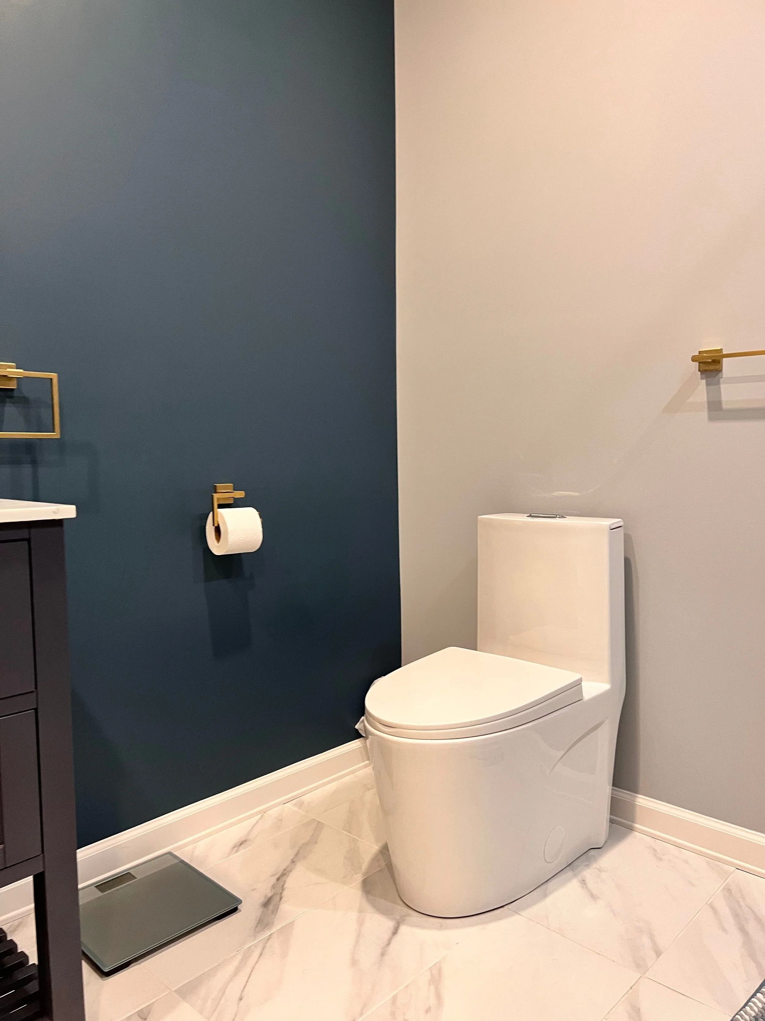 Modern bathroom with a white toilet, a dark blue accent wall, a gray scale on the floor, gold towel racks, and a white countertop with a sink.