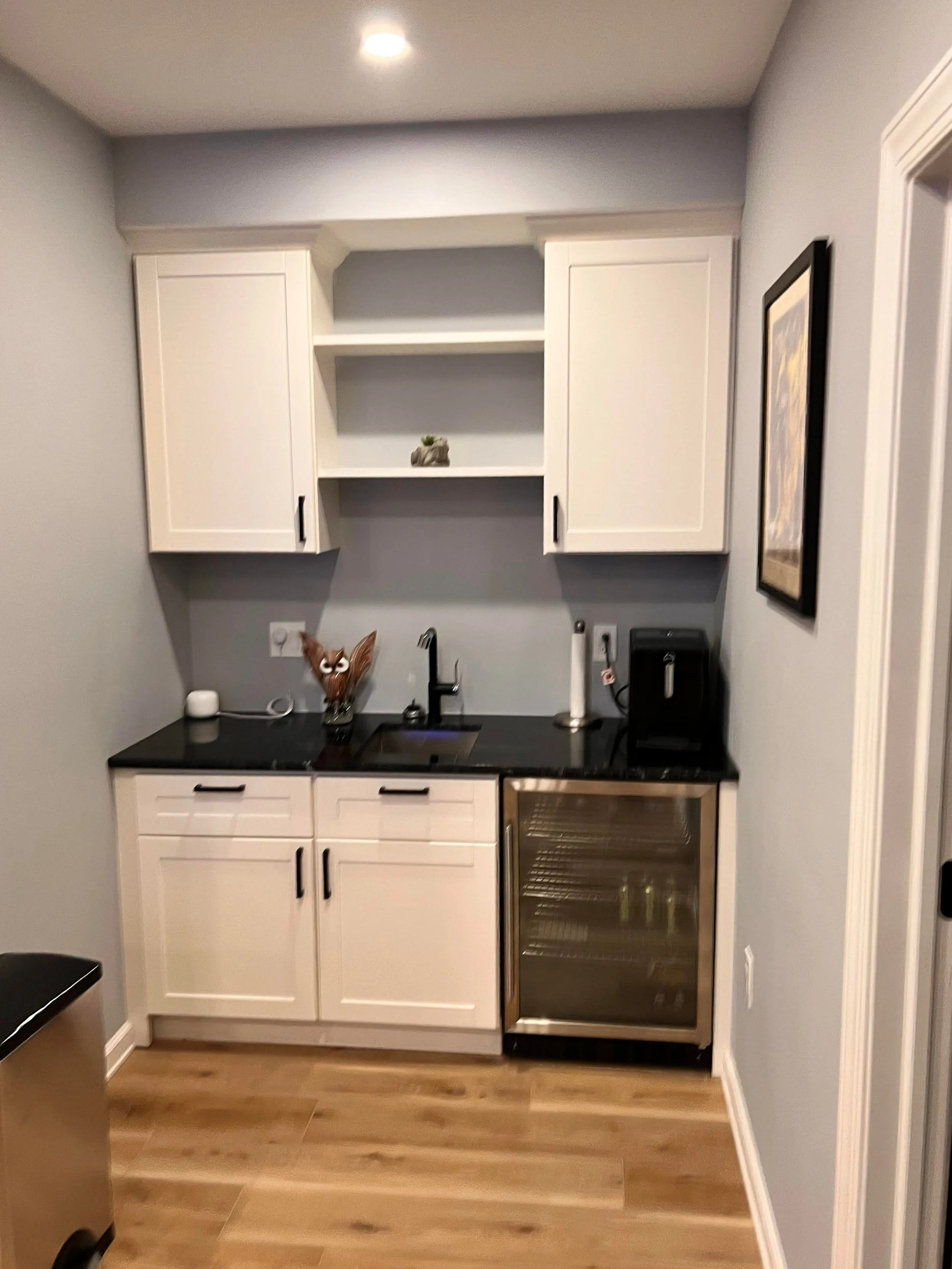 Small kitchenette with white cabinets, black countertop, a small wine fridge, and a few small appliances, decorated with a framed picture on the wall.