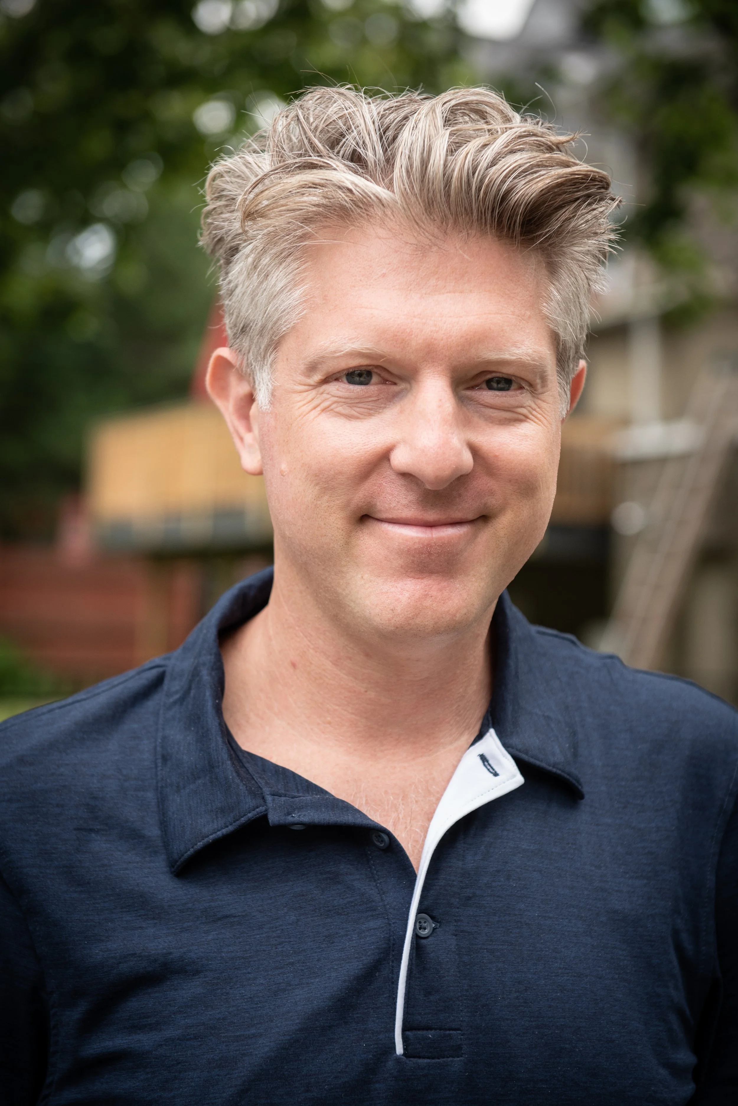 Man with gray hair smiling outdoors wearing a blue collared shirt.
