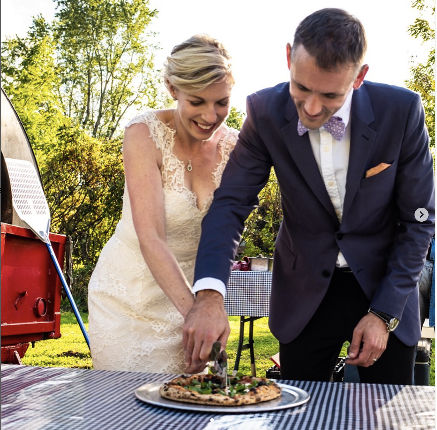 Couple dressed in wedding attire slicing a pizza outdoors during a celebration with trees in the background.