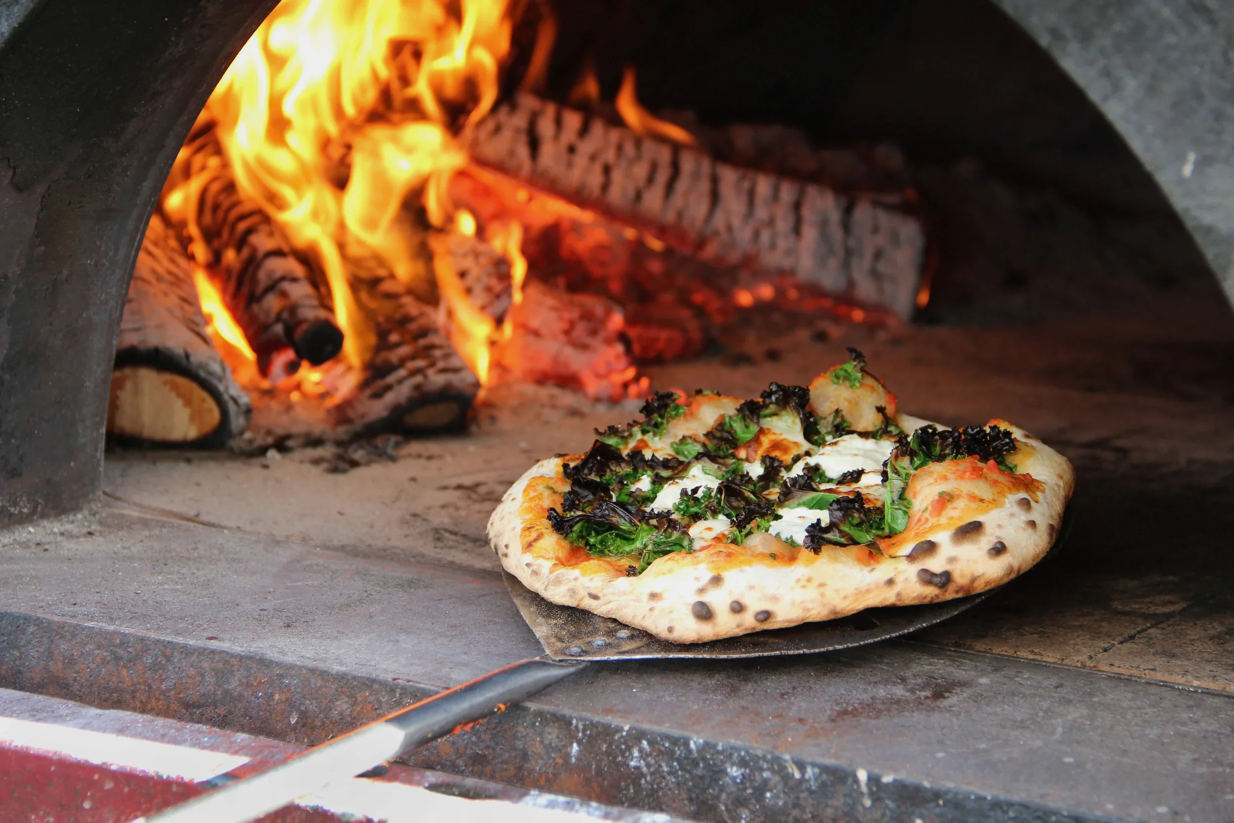 Pizza baking in a wood-fired oven with flames visible in the background.