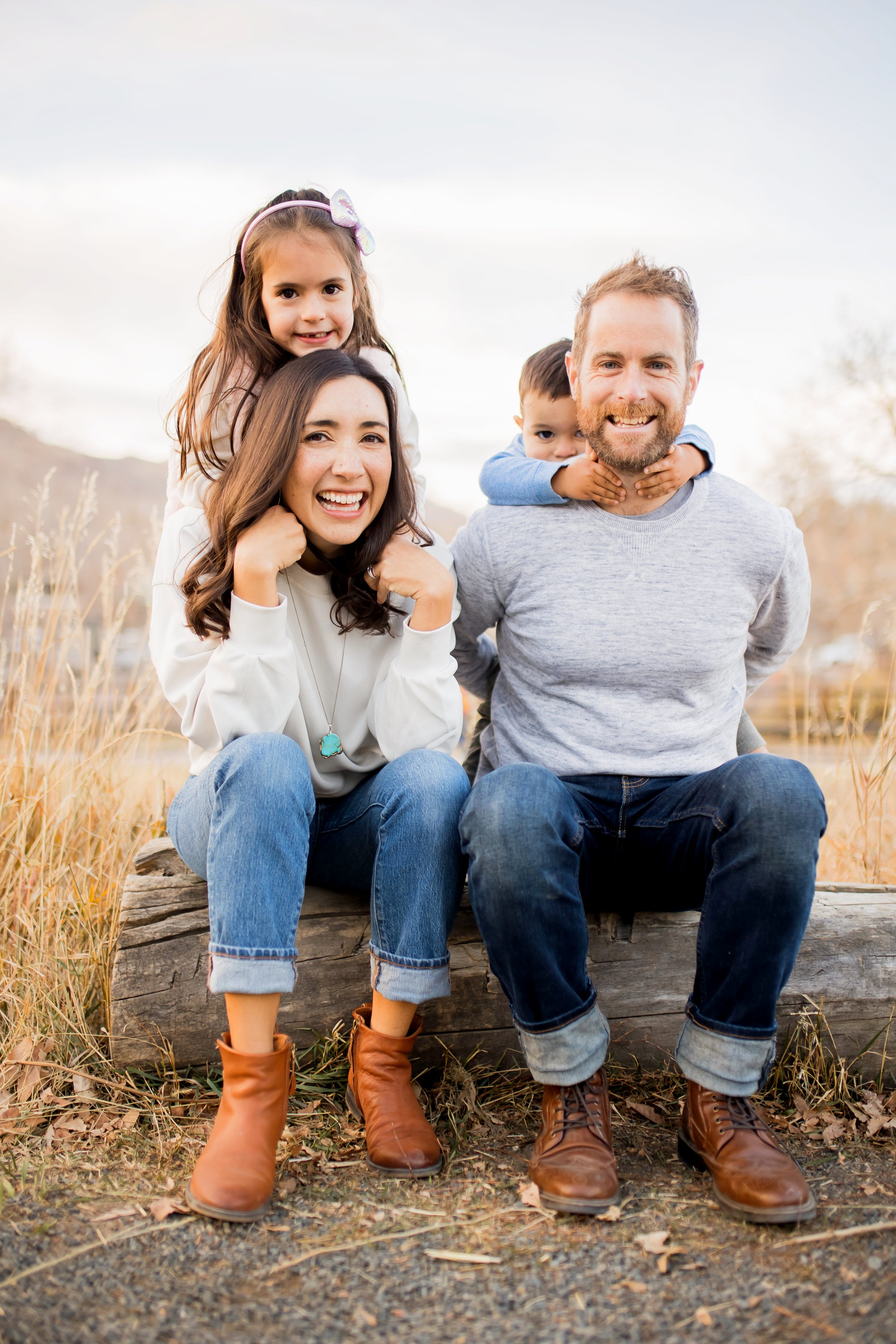 Family of four sitting on a log in an outdoor setting with tall grass, smiling at the camera. The mother is in front with her daughter on her shoulders, and the father with his son on his back, all dressed casually.