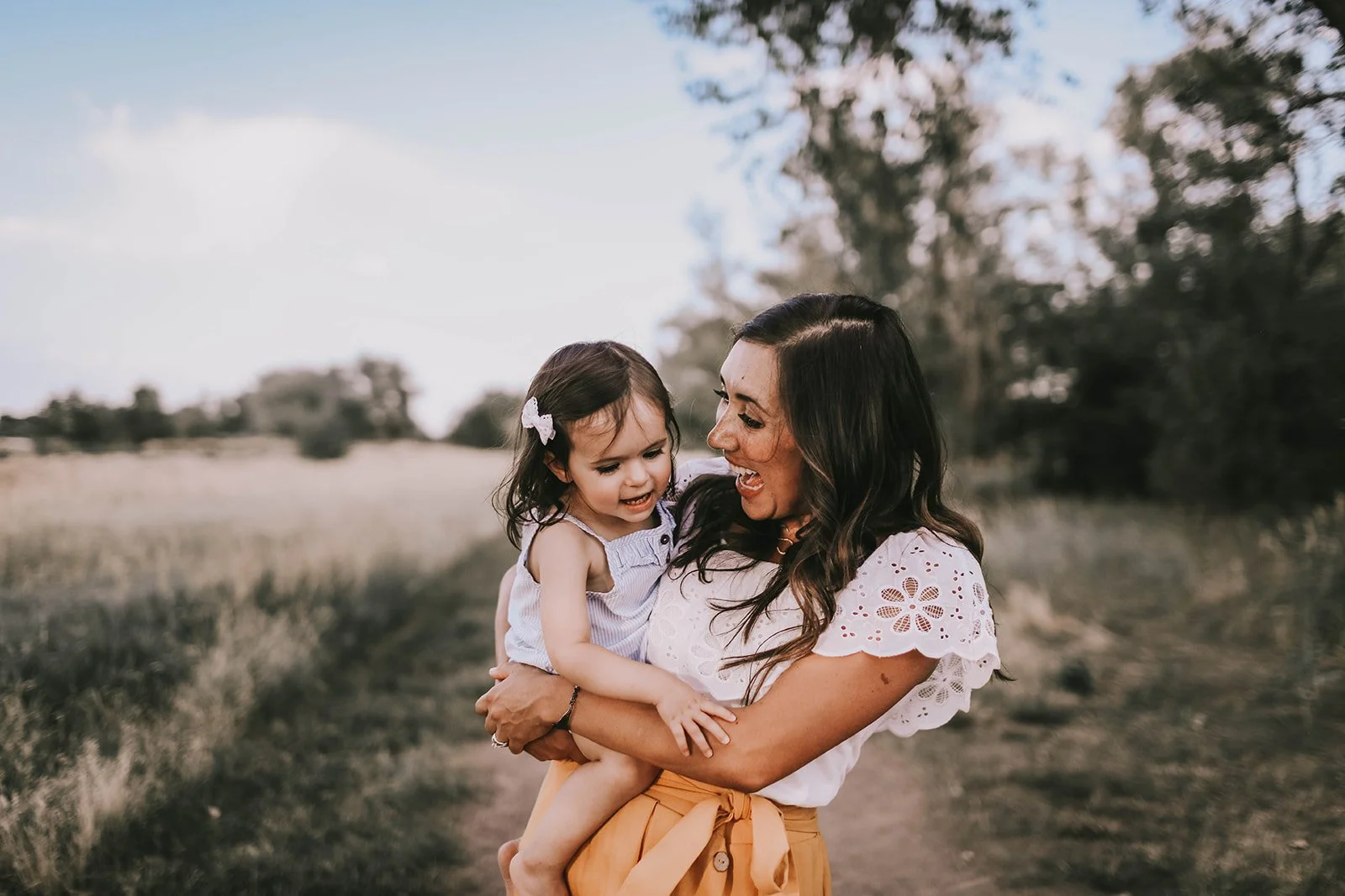 A woman holding a young girl outdoors in a natural setting, both smiling and looking at each other.