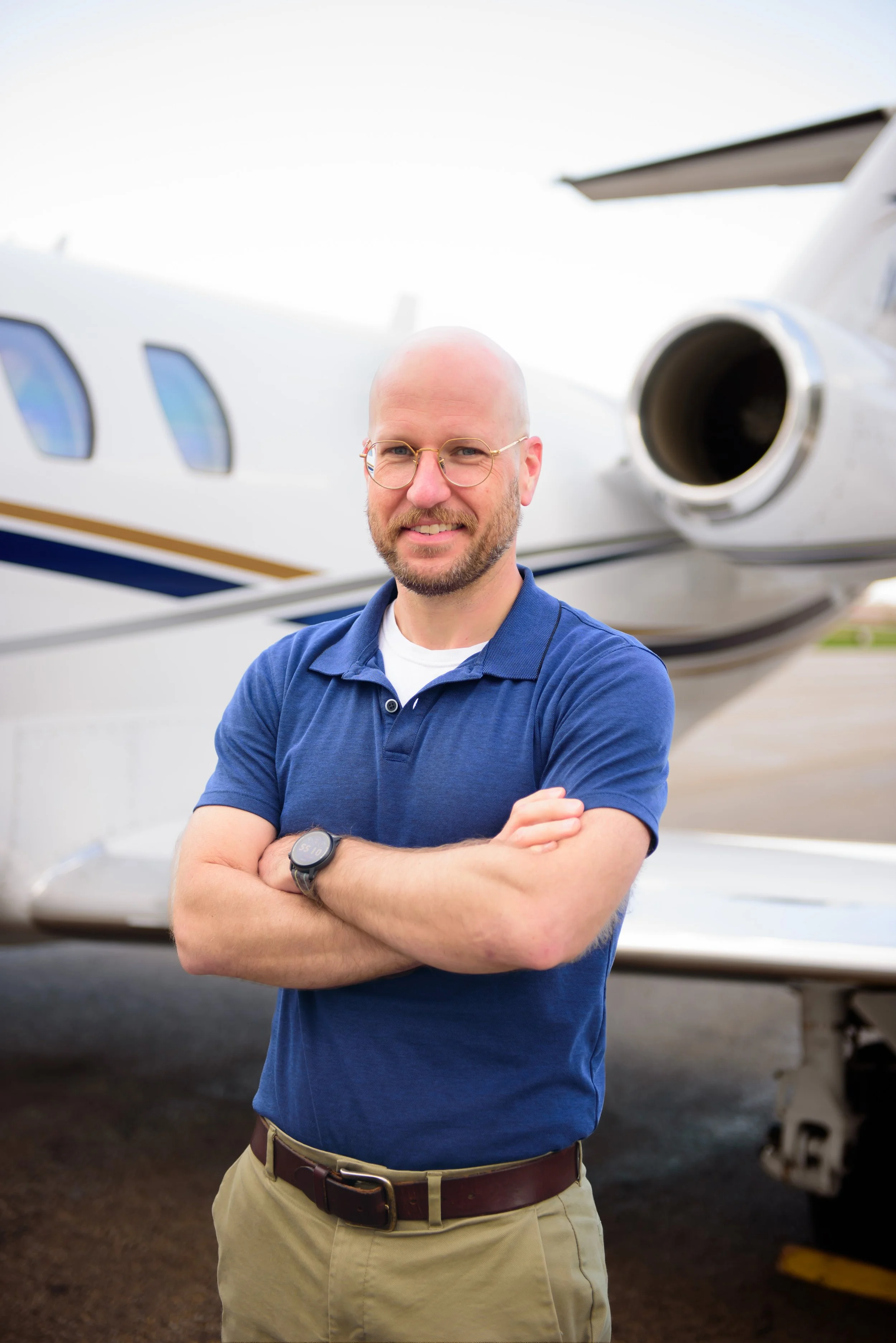 Portrait of a bald man with glasses, beard, and mustache, smiling with arms crossed, standing in front of a small private jet.