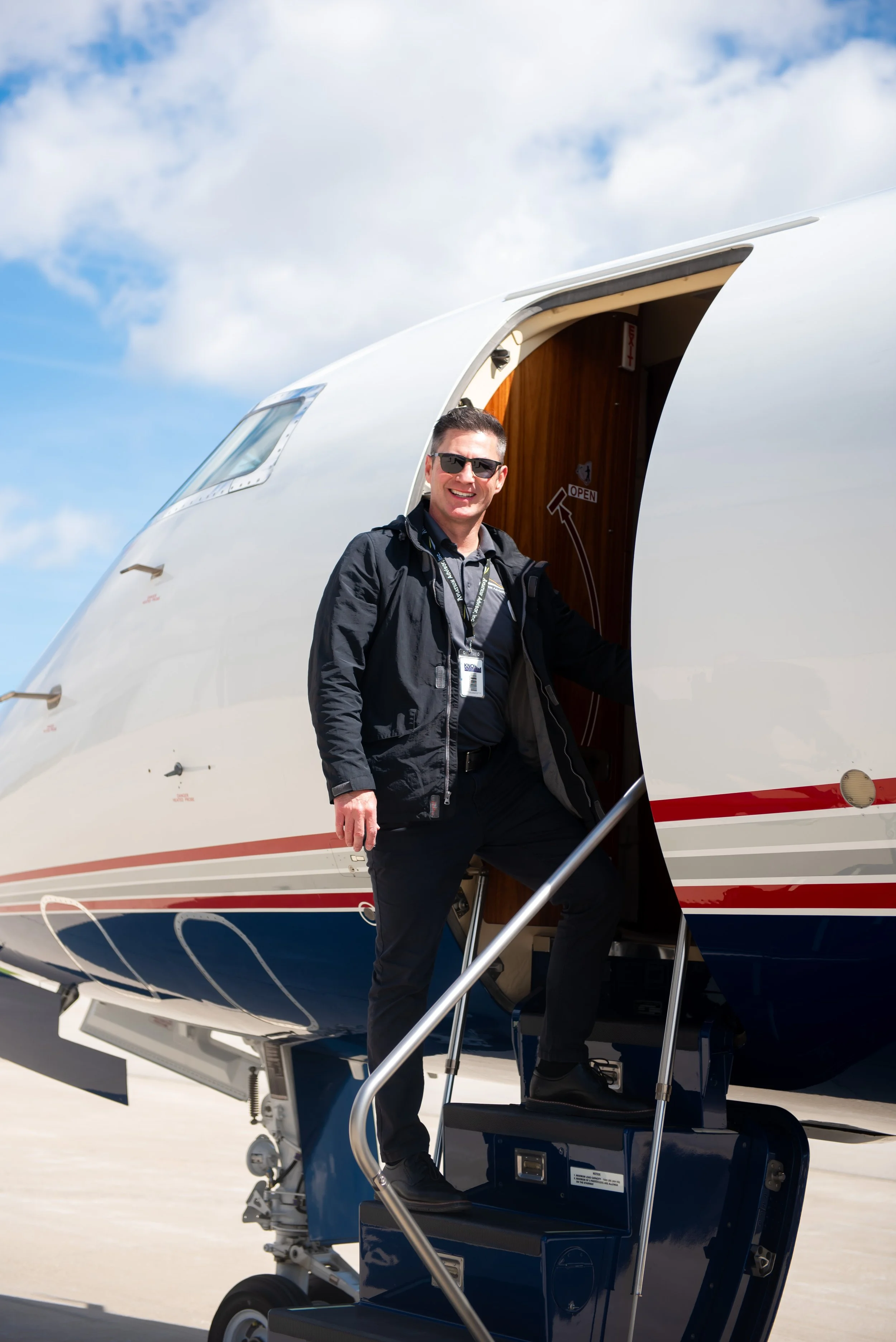 A man wearing sunglasses and a black jacket steps onto a private jet using a small stairway, with the sky and clouds in the background.
