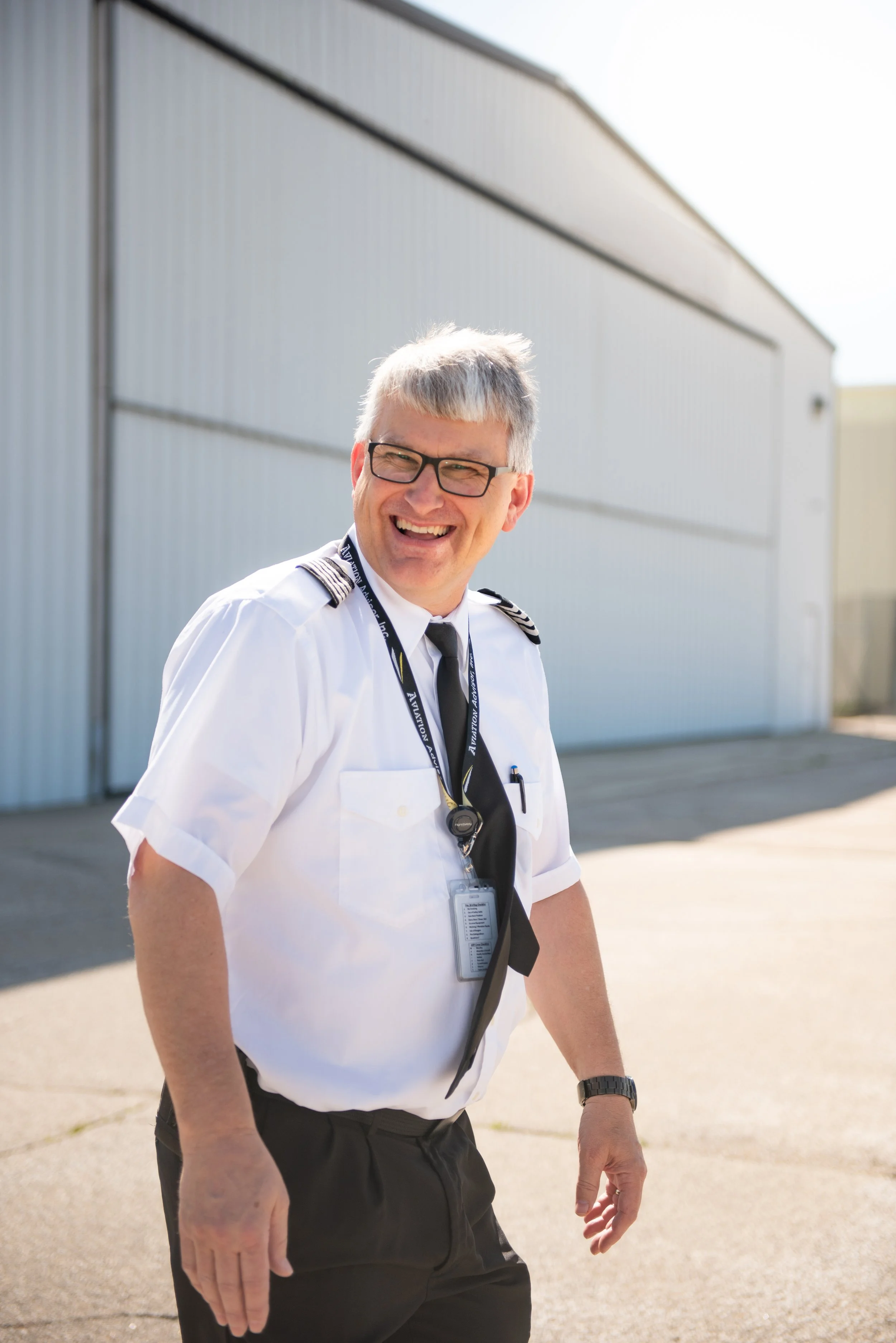 A smiling man in a pilot uniform standing outdoors near a large industrial building.